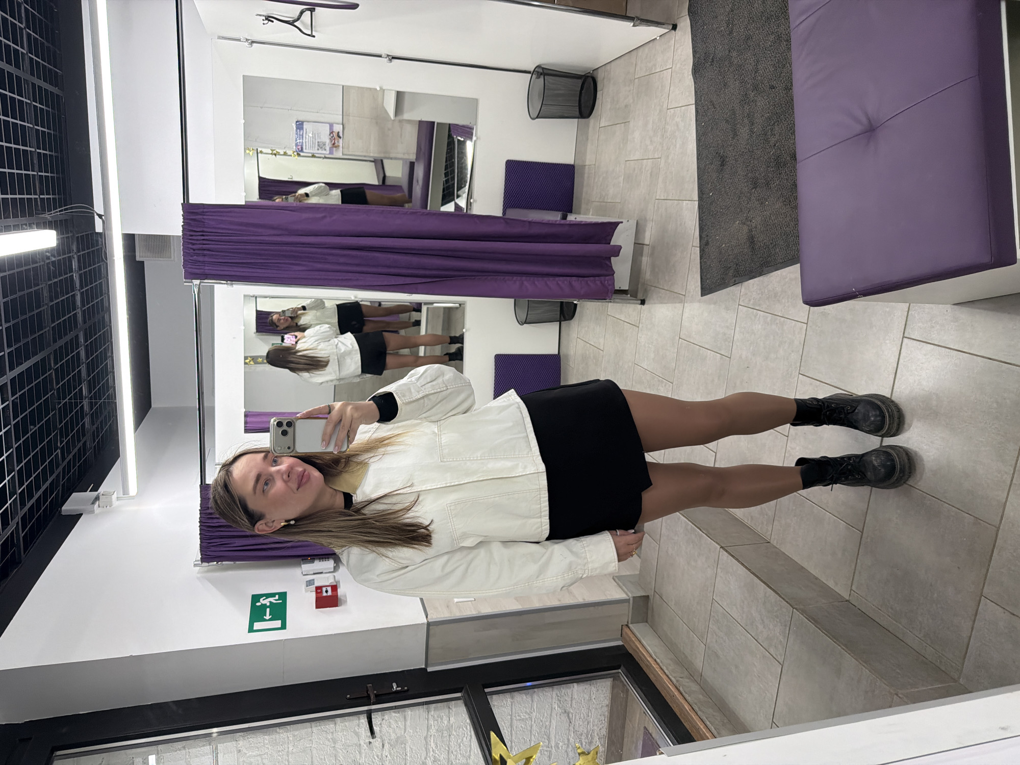 A young woman in a white jacket and black skirt is taking a selfie in a fitting room with purple curtains and seating. She is wearing black boots. The fitting room is in Moscow, Russia. The room has a grid ceiling and tiled flooring.