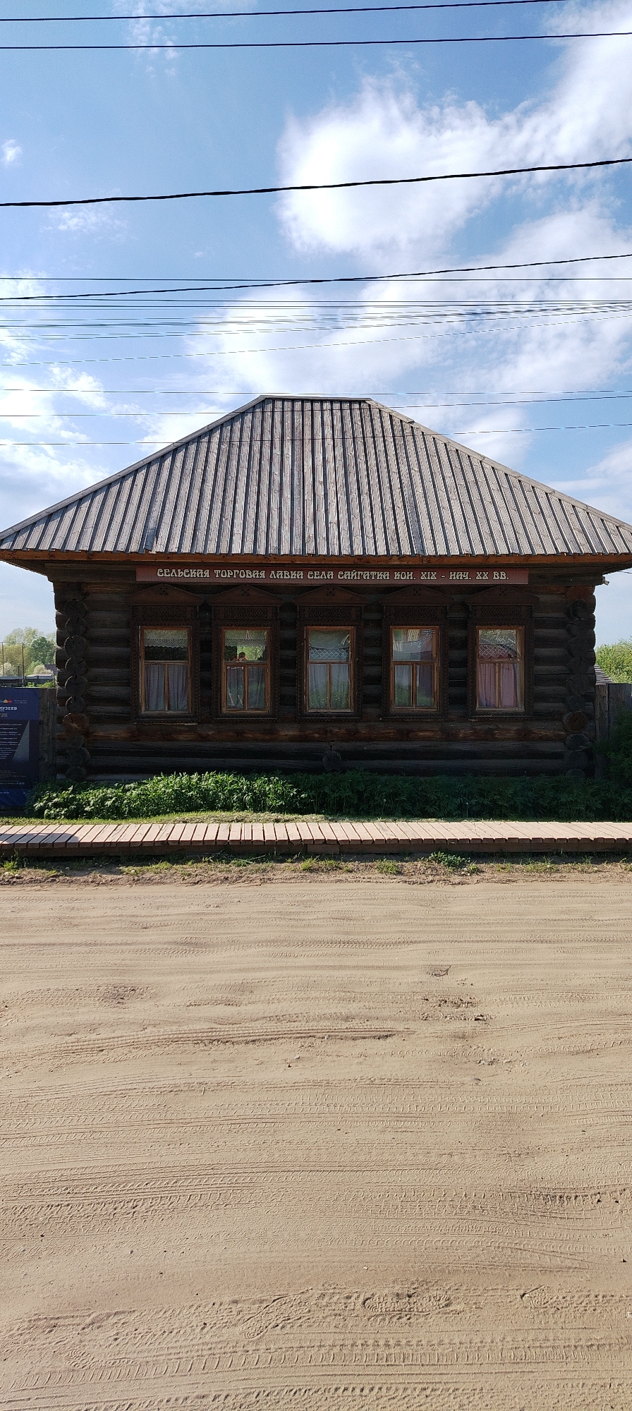 The image shows a wooden building in what appears to be the city of Chaikovsky, Russia. The building is rectangular, made of dark logs, and has a gray, gabled roof with vertical planks. Five windows are visible on the front facade, each framed with decorative wood carvings and with curtains or coverings inside. A sign above the windows is inscribed in Cyrillic text: "СЕЛЬСКАЯ ТОРГОВАЯ ЛАВКА СЕЛА САЙГАТКА кон. XIX - нач. XX вв." suggesting the building served as a village shop in the late 19th to early 20th centuries. A low wooden walkway runs in front of the building, with green vegetation growing beneath. In the foreground, the ground is bare dirt, marked with tire tracks. Power lines stretch across the upper portion of the image against a backdrop of a blue sky with scattered white clouds. The overall scene appears to be a museum or preserved historic site.