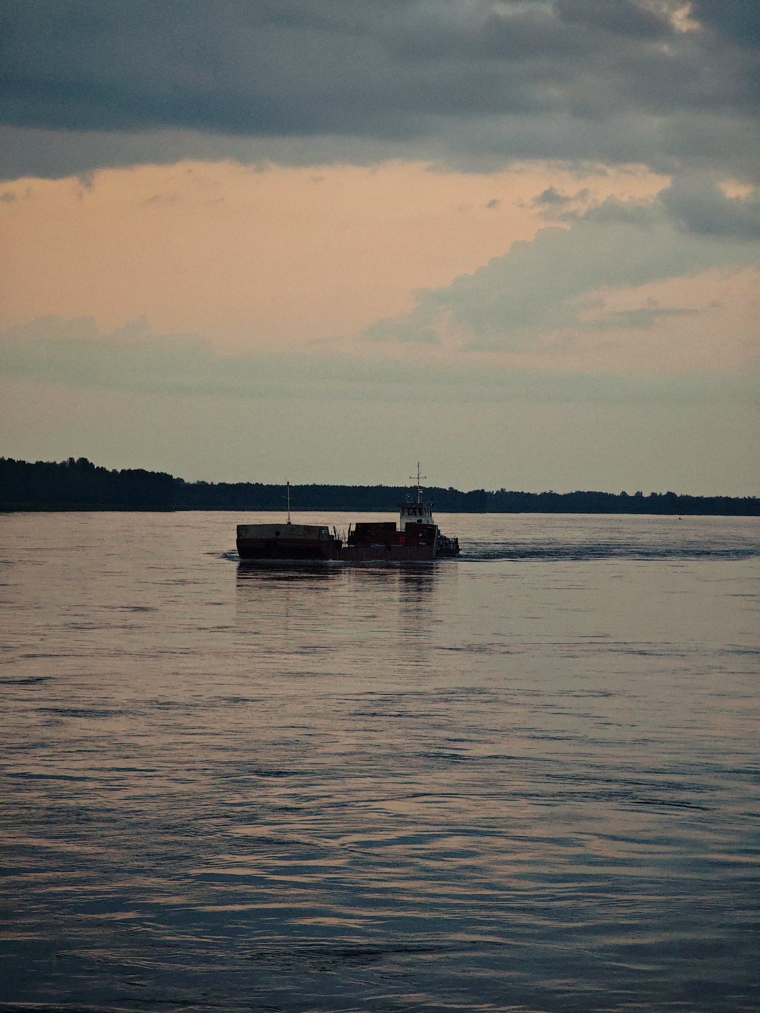 A barge is traveling on a wide river, moving from left to right in the frame. The barge appears to be hauling cargo. The water has a rippling surface, reflecting the muted colors of the sky. In the background, a dark line of trees forms the horizon. The sky above is filled with clouds, with shades of gray and pale pink. The scene suggests either sunrise or sunset. The location is identified as the city of Kazachinskoe, Russia.