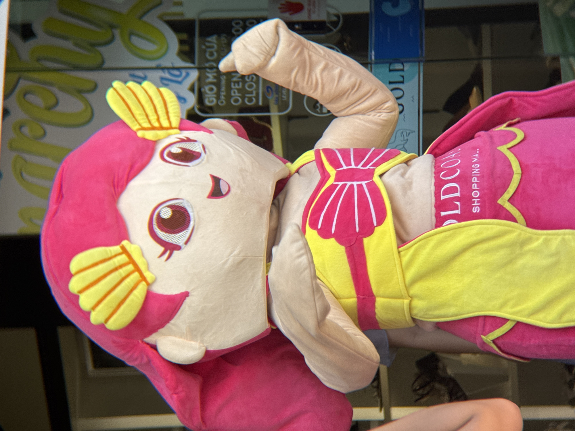 An outdoor, close-up, eye-level, medium shot shows a person in a mermaid costume with bright pink hair and yellow shell-shaped accessories on their head. The costume is predominantly pink and yellow, featuring a bow on the chest and a yellow sash. The character has large, stylized eyes and an open mouth with a smiling expression. The right arm of the costume is raised, with the thumb extended upwards in a "thumbs up" gesture.
In the background, a glass storefront displays signage. One sign, visible at the top of the image, has large yellow cursive lettering, partially obscured, that appears to say "Mercy." Below this, another sign is written in Vietnamese and English, indicating "GIỜ MỞ CỬA" (Opening Hours) and "OPEN" followed by numbers. Another sign below reads "CLOSING" and more numbers. The name "GOLD COAST SHOPPING MALL" is visible in white lettering on a blue sign in the lower portion of the image. The reflection in the glass suggests an urban environment.
The image appears to have been taken in Nha Trang, Vietnam, given the presence of Vietnamese text on the signage and the general visual context of a commercial area. The lighting suggests it is daytime.