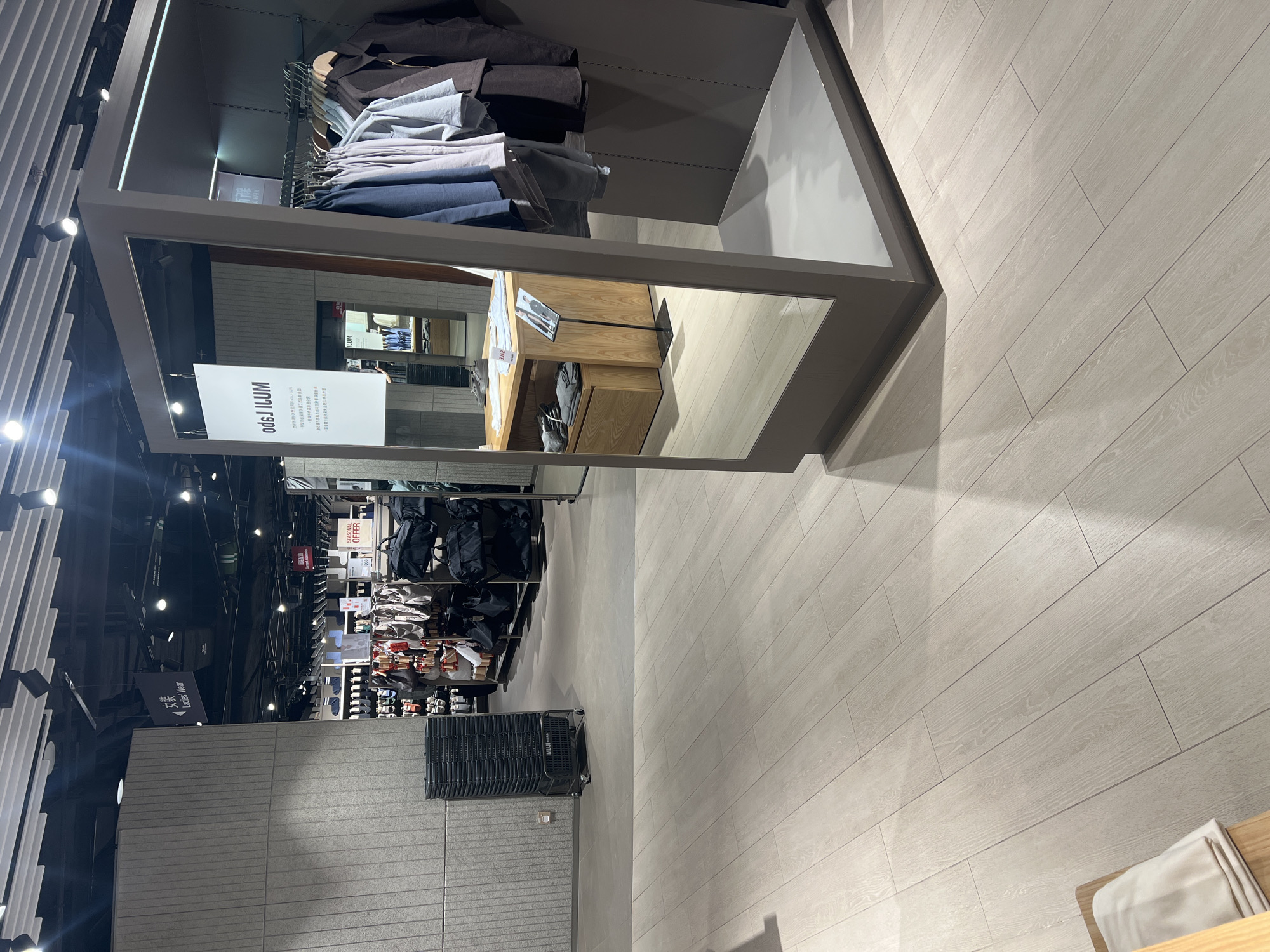 The image shows the interior of a Muji store in Hong Kong, China. The store features a clean and minimalist design with light wood flooring and grey walls. In the foreground, a large mirror is placed on a grey frame, reflecting the surrounding environment. Clothing racks filled with Muji apparel are visible to the right of the mirror. In the background, there are displays of clothing and accessories, as well as signage indicating different sections of the store, such as "Ladies' Wear." A stack of black shopping baskets is positioned near the left wall. The lighting is bright and even, typical of a retail environment.