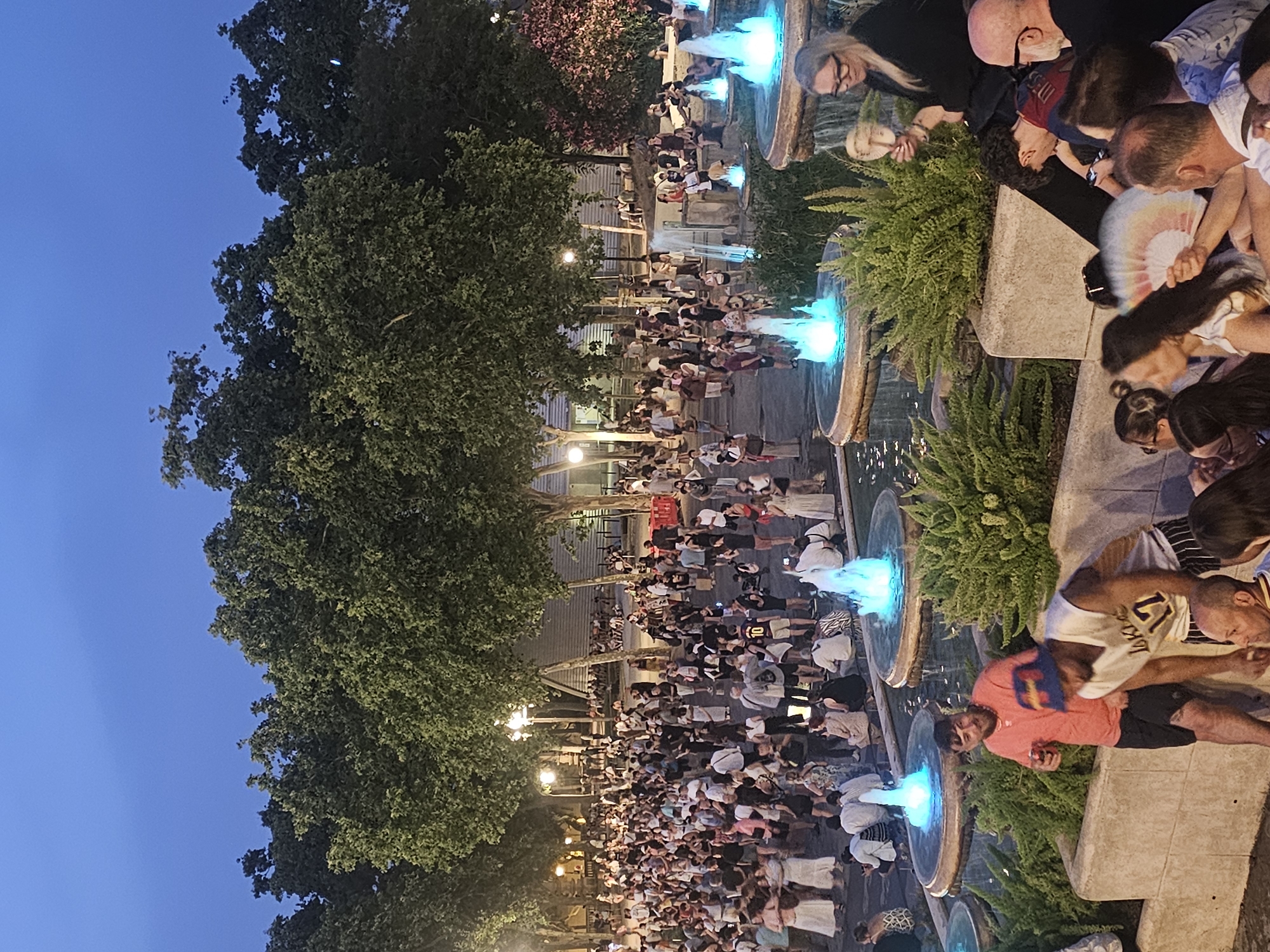 This is a nighttime photograph of a crowded public square, likely taken in Barcelona, Spain. The scene features a large group of people gathered around illuminated fountains. The fountains emit a bright blue light, creating a striking visual against the twilight sky and the dark green foliage of trees bordering the square. Many people are standing or sitting, some observing the fountains, others conversing. Several individuals are holding drinks and some are fanning themselves. The architecture of the surrounding buildings is visible in the background, with lampposts providing additional illumination. The overall atmosphere appears to be festive and social, suggesting an evening event or gathering.