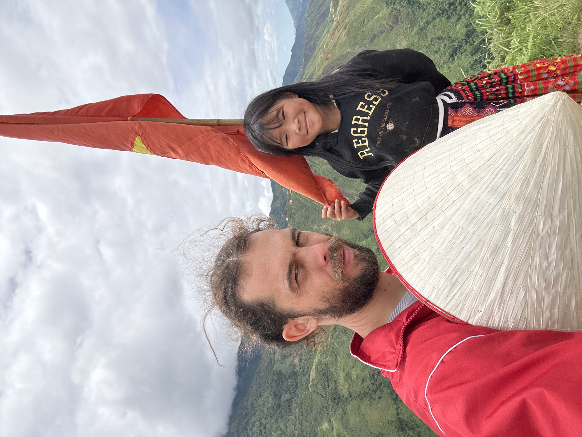 In Ta Xua, Vietnam, a man with a beard and tied-back curly hair, wearing a red jacket with white trim, poses for a selfie. He holds a large, conical straw hat in front of him. Next to him, a young girl smiles at the camera. She has dark hair and wears a black hooded sweatshirt with "REGRESS" and "PASS ON THE CLASS-CS" printed on it, along with a colorful, patterned skirt. Behind them, a red flag with a yellow star, likely the Vietnamese flag, flies from a bamboo pole against a backdrop of dramatic, cloud-covered mountains and lush green valleys. The environment appears to be a scenic viewpoint in the mountainous region.