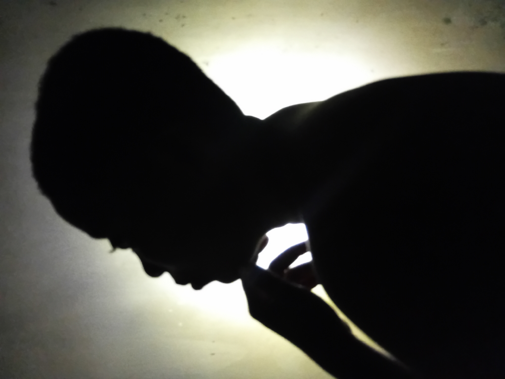 The image shows a silhouette of a person with their head bowed and hand near their face. The background is a diffuse light source, possibly a window or a light fixture. The location and city are unknown.
