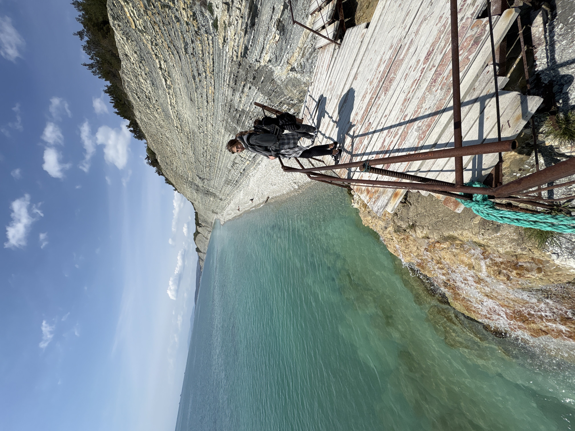 This image captures a dramatic coastal scene in Gelendzhik, Russia, showcasing a person standing on a narrow walkway built along a towering cliff face.
The central subject is a female individual, seen from behind and slightly to her left, standing on a wooden walkway. She is dressed in a plaid shirt or jacket, dark leggings, and a gray scarf, with a dark backpack on her shoulders and sunglasses resting on her head. Her hands rest on a rusty metal railing that forms part of the walkway's barrier.
The walkway itself is constructed from weathered, light-colored wooden planks, some showing signs of wear and peeling paint. It features sturdy, albeit rusty, metal railings that extend downwards towards the water. A thick, braided green rope is visibly secured to the lower portion of one of these railings.
To the left of the walkway rises a massive, stratified cliff, characterized by distinct horizontal layers of light grey and beige rock. The very top of the cliff is covered with a line of dense green trees. Below the walkway and cliff, the clear, turquoise waters of the sea gently lap against a narrow, rocky and pebbled shoreline. The water appears calm, with small whitecaps visible where it meets the shore. In the distance, the vast expanse of the sea meets the horizon under a bright blue sky dotted with scattered white clouds.
The scene suggests a bright, sunny daytime, evidenced by the sharp shadows cast by the person and the railings on the wooden planks. The overall impression is one of a breathtaking natural landscape combined with a man-made path offering stunning views. No visible text is present in the image.