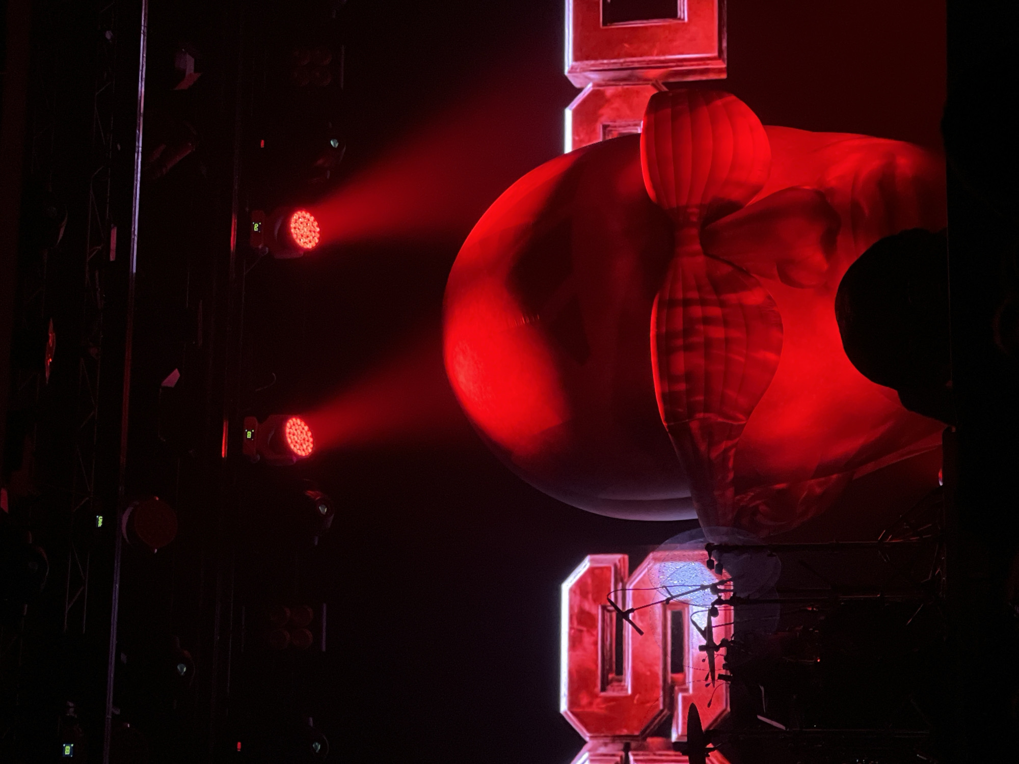 The image, likely taken at a concert or performance venue, presents a dark scene illuminated by red lighting. On the left, several stage lights are visible, mounted on a dark metal frame. A beam of red light emanates from one of these lights, directed toward the center of the image.
In the center, a large, illuminated object resembling an inflated figure is positioned next to large letters. This creates a surreal effect, with the lighting highlighting the form and texture of the figure.
On the right, a dark figure, possibly a person or object, is partially silhouetted against the red light.
Based on available information and visual cues, a specific location within the city of Kazan, Russia, cannot be definitively determined.