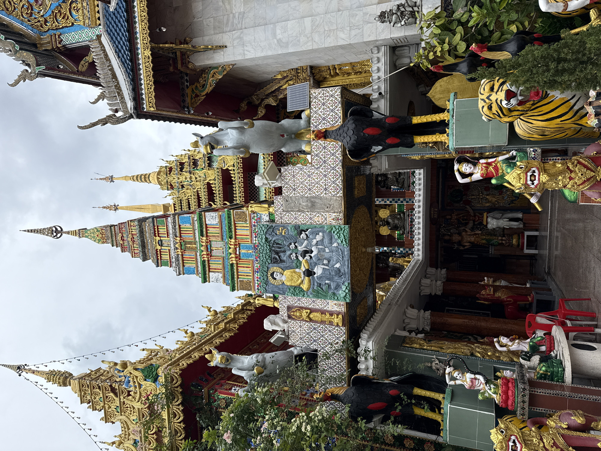 The image shows the ornate facade of Wat Samphran, a Buddhist temple located in Nakhon Pathom Province, Thailand, approximately 40 kilometers west of Bangkok. The temple is known for its towering, dragon-wrapped cylindrical structure, though this specific image focuses on the main entrance area.
The central feature is a multi-tiered, colorful spire structure adorned with gold, green, blue, and red tiled patterns, topped with a golden finial. Flanking this central spire are two other golden spires. Below the main spire, a bas-relief carving depicts Buddha in meditation surrounded by disciples.
Numerous decorative elements are present:
* Two large, white statues of rearing horses with golden accents are positioned on ledges.
* Two large, black rooster statues with red markings and yellow legs stand prominently in the foreground.
* Various smaller statues are visible, including figures of women in traditional attire, mythological creatures (such as a golden dragon-headed figure and a tiger), and what appear to be seated Buddha images.
* The facade is elaborately decorated with gold trim, colorful mosaic tiles, and intricate carvings.
* A solar panel is attached to the wall behind the right-hand horse statue.
* Greenery, including flowering plants and a small coniferous tree, is interspersed with the statues and architectural elements.
* The sky above is overcast.
* A red plastic chair is visible near the center-left of the image.
The overall impression is one of vibrant religious and artistic expression, characteristic of Thai temple architecture.