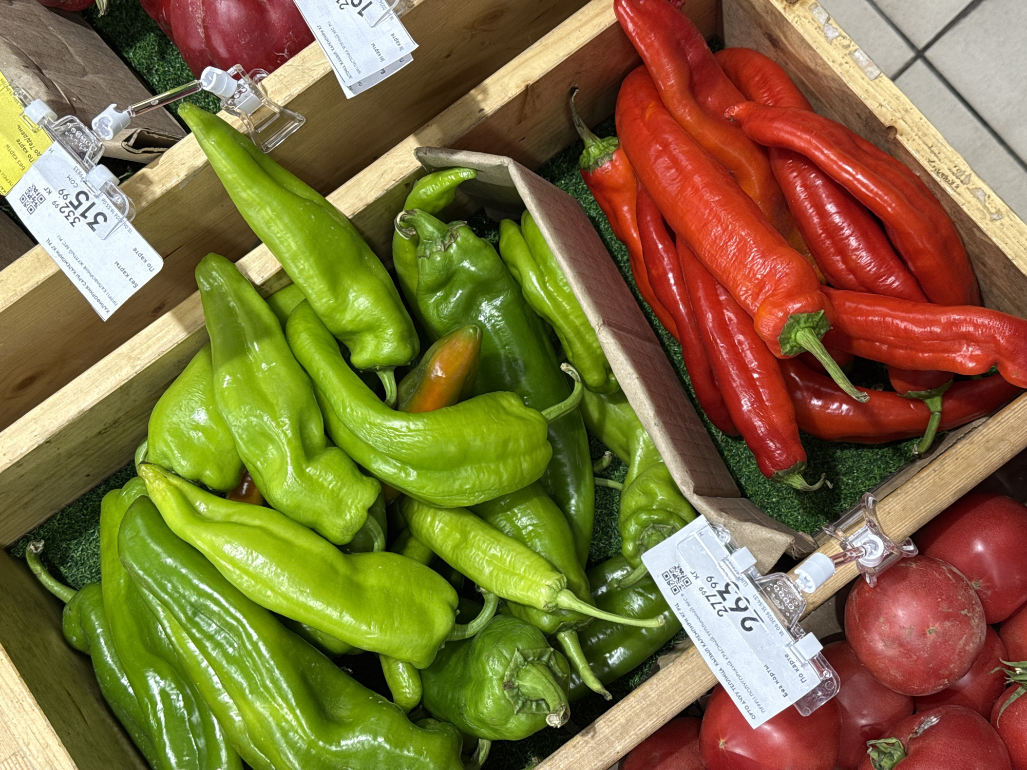 A top-down, angled close-up shot shows a display of fresh produce at a market. In the foreground, a wooden crate is filled with bright red chili peppers. Behind them, another wooden crate is filled with numerous long, bright green bell peppers. The green peppers are packed tightly together and spill over the sides of their crate. To the left, a pile of round, red tomatoes is visible. Several price tags with Cyrillic text and barcodes are attached to the wooden crates and the display. The environment appears to be a grocery store or farmer's market, with hints of shelving and a tiled floor visible in the background. The lighting is bright and even, highlighting the vibrant colors of the produce.