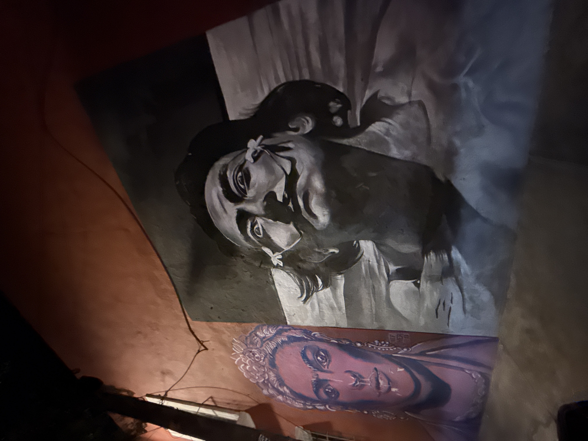 The image shows two adjacent murals on a wall in Puebla, Mexico. The top mural is grayscale, depicting a woman with a flower near her eye, set against a light background with dark accents. The lower mural is in color, showing a face with decorative elements around the hair, rendered in shades of pink and purple. The surrounding wall is orange. The photograph is taken at a low angle, indicating the mural is located at a height, with dark areas in the foreground suggesting a dimly lit environment.