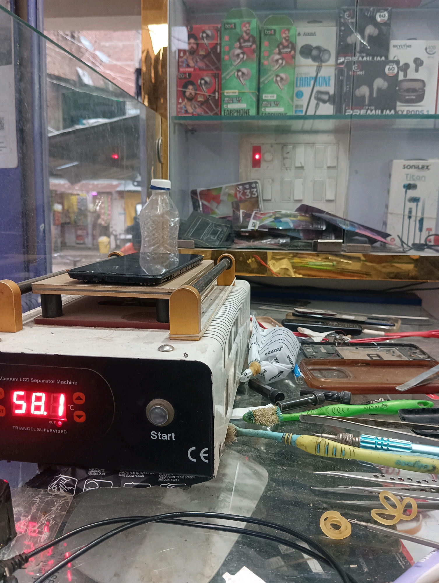 The image depicts a close-up view of a mobile phone repair shop's workbench in Dakhin Parsa, India. The foreground is dominated by a "Vacuum LCD Separator Machine" with a digital display showing "58.1 C" and the word "OUT" illuminated in red. A black smartphone is positioned on top of the machine, secured by a clear plastic bottle filled with water acting as a weight.
The workbench surface is cluttered with various tools and materials related to phone repair. These include tweezers, screwdrivers, brushes, a brown phone case, and several rubber bands. In the background, shelves are visible, stocked with electronic accessories such as earphones and phone cases. A power switchboard with a red indicator light is also present on a shelf. The overall environment appears to be indoors, likely within a small commercial establishment. No individuals are visible in the frame.