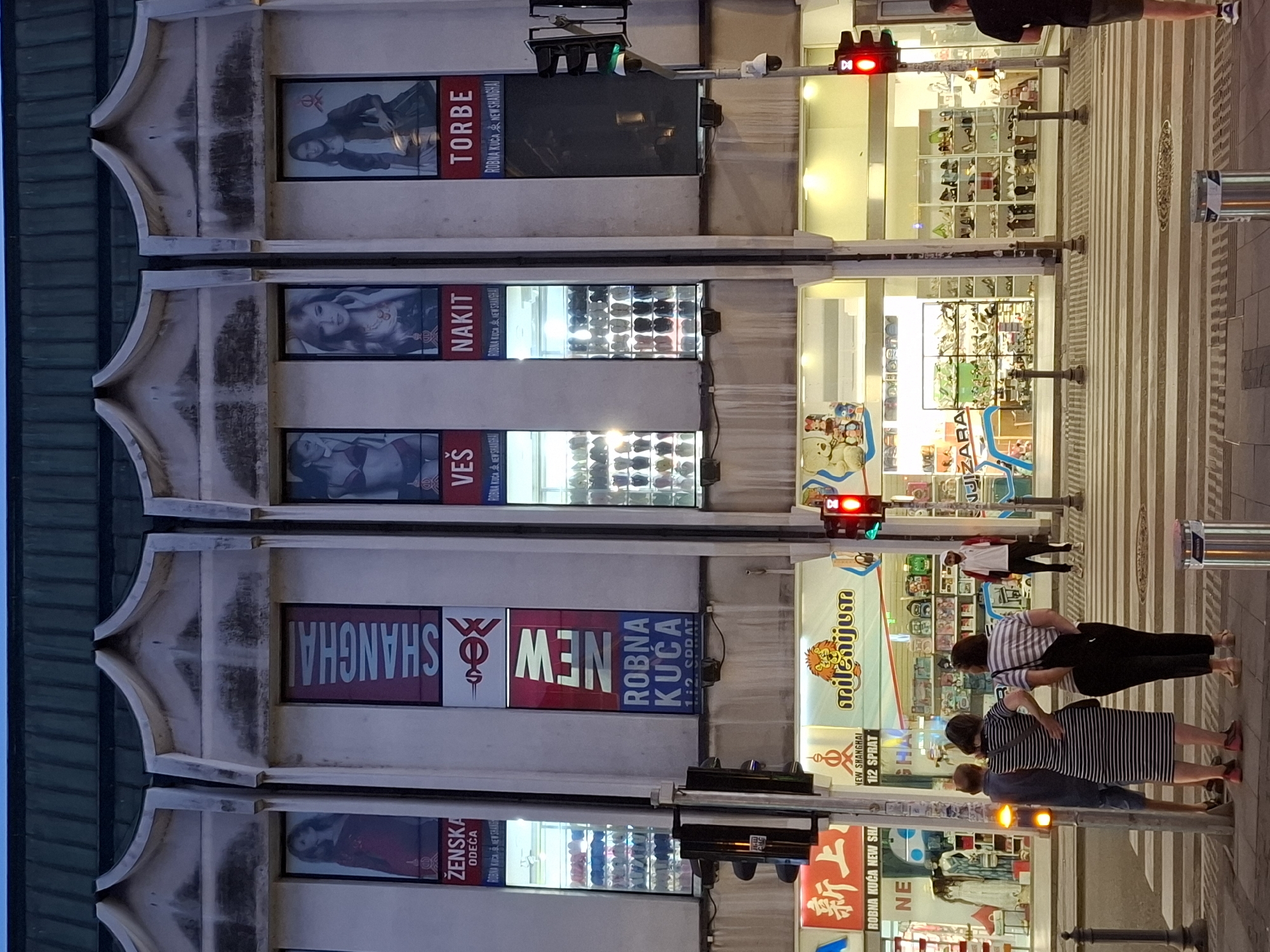 The image shows a street scene in Zemun Urban Municipality, Serbia, likely in the evening or dusk. In the background, a large, multi-story building with a dark green roof dominates the scene. The building's facade is a light-colored concrete with distinct architectural features.
The upper floors of the building feature large rectangular windows displaying advertisements. From left to right, the advertisements appear to be for:
1. "ŽENSKA ODECA" (Women's Clothing) with an image of a woman in a red dress.
2. "SHANGHAI NEW" and "ROBNA KUCA" (Department Store) with a stylized logo.
3. "VEŠ" (Underwear) with an image of a woman in lingerie.
4. "NAKIT" (Jewelry) with an image of a woman wearing jewelry.
5. "TORBE" (Bags) with an image of a woman holding a bag.
Below these advertisements, the ground floor of the building appears to be a commercial space with large glass windows revealing shops. Various store signs are visible, including "ROBNA KUĆA NEW SHANGHAI," "milenijum," and "ZARA." The shops display clothing, shoes, and other merchandise.
In the foreground, a pedestrian crossing is visible, with several people waiting to cross. Three individuals are standing on the left side of the crossing: a man wearing a blue shirt and shorts, a woman in a black and white striped dress, and another woman in a black and white striped top and dark pants. They are facing away from the camera. A man in a white polo shirt and dark pants is standing on the right side of the crossing, looking towards the shops.
Traffic lights are prominent in the scene. A red traffic light is visible on the right side of the image, and a traffic signal with a yellow light is on the left. A security camera is mounted on a pole near the traffic signal on the right. Street lamps and lights from the shops illuminate the area. The overall atmosphere suggests a commercial street in an urban setting.