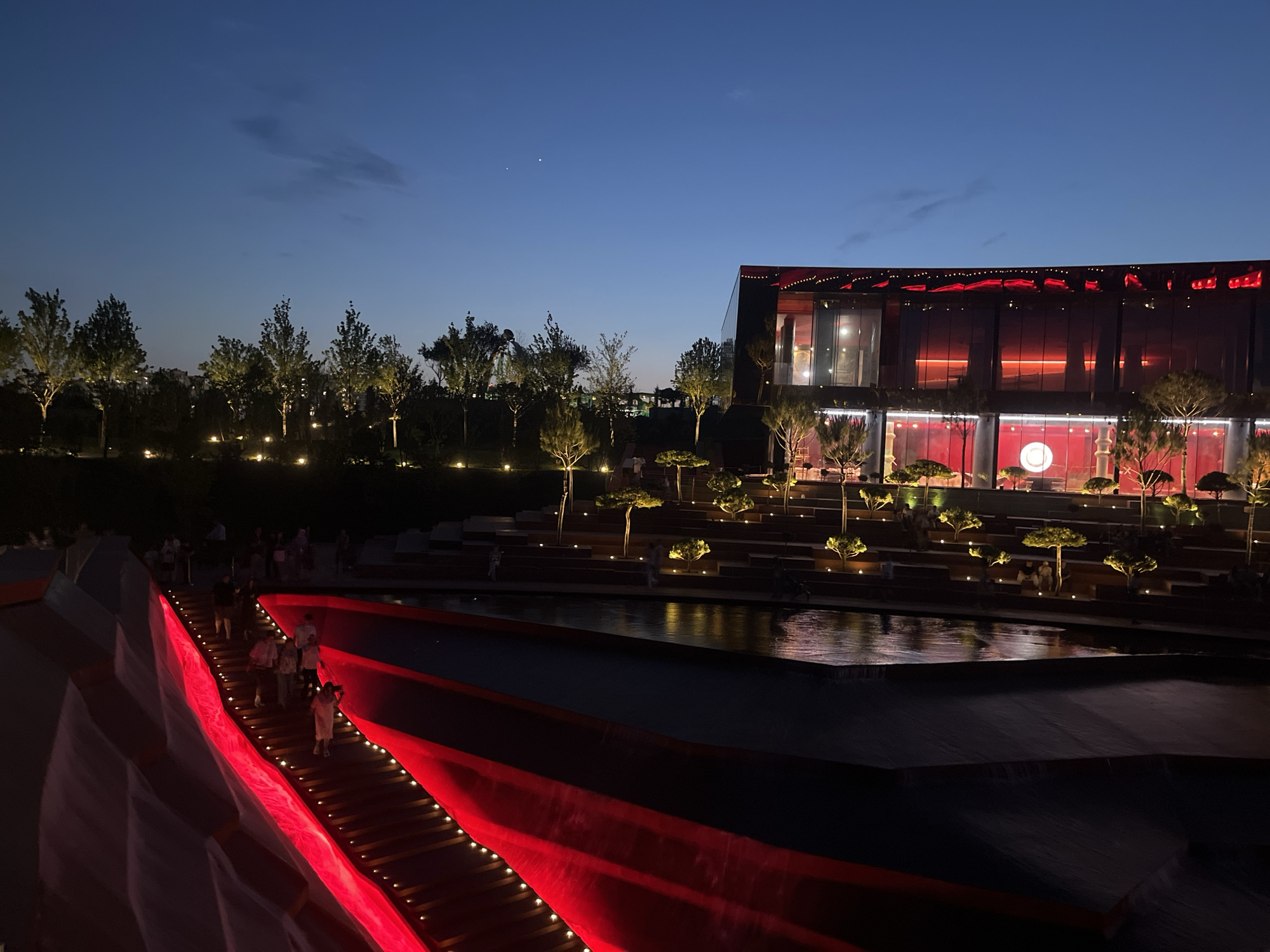 The image depicts an outdoor scene at dusk or early evening in Krasnodar, Russia. The sky is a deep blue, transitioning from twilight. A modern, multi-story building with a dark facade and extensive glass windows dominates the background. The building is illuminated with vibrant red lights along its roofline and interior, reflecting on the glass.
In the foreground, a wide staircase or ramp is dramatically lit with bright red lights along its edges and stair risers, creating a striking visual contrast. Several people are walking up this illuminated pathway. The setting appears to be a park or public space, with tiered landscaping, small, sculpted trees, and numerous small, warm-toned lights illuminating the pathways and gardens. A body of water, possibly a reflecting pool or a series of cascading water features, is visible in the mid-ground, reflecting the ambient light.
The overall atmosphere is one of an evening gathering or event, with the modern architecture and dynamic lighting suggesting a contemporary urban entertainment or cultural space. The presence of people suggests activity, possibly a promenade or attending an event. The clear sky and gentle lighting indicate fair weather. There is no visible text in the image.