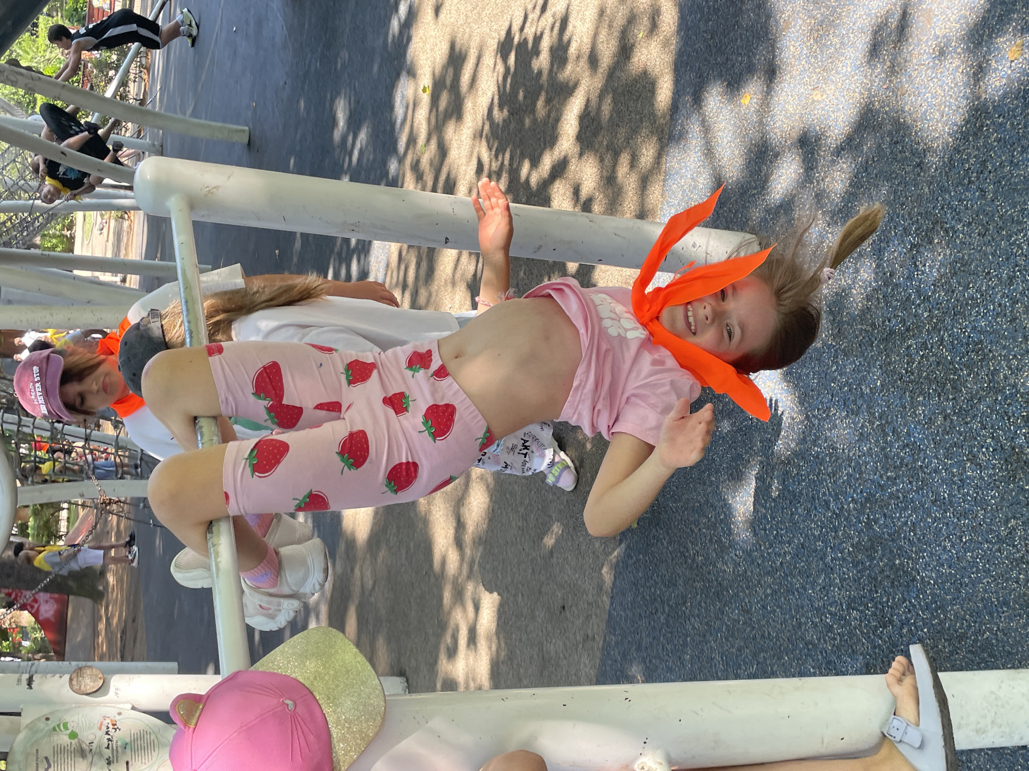 An outdoor daytime shot shows a young girl hanging upside down from a metal bar at a playground in Sochi, Russia. The girl, with blonde hair in a ponytail, is wearing a pink t-shirt with strawberry patterns and pink shorts. She has an orange bandana tied around her neck. Her face is towards the camera and she is smiling. Her legs are bent at the knees and her feet are resting on the metal bar. In the background, there are other children playing on playground equipment. The ground is covered with a dark blue rubber surface.