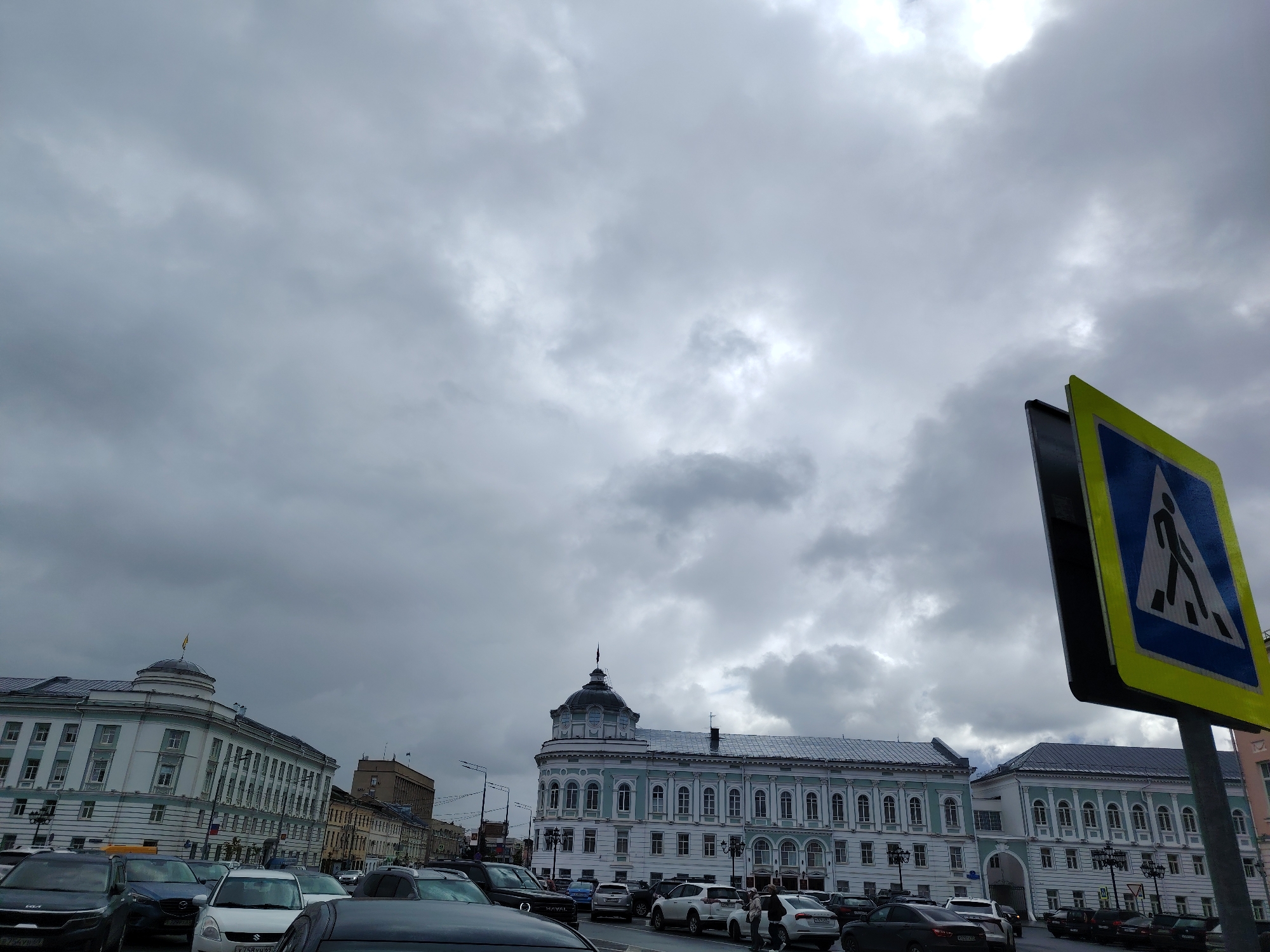 A panoramic view captures a city street in Tver, Russia, under a heavily overcast sky. In the foreground, a pedestrian crossing sign with a blue triangle and a black silhouette of a person walking stands prominently on the right. The street is lined with several cars, indicating moderate traffic. On the left side of the street, a grand, light-colored building with classical architectural features dominates the scene. This building has numerous windows and a rounded section on its left end, topped with a dome-like structure. Further down the street, another similarly styled building, painted in white and light blue, with a prominent dark dome, is visible. The background features more multi-story buildings, some with flags on top, and a cloudy sky that suggests a cool or potentially rainy day. The scene appears to be an urban setting, likely a main thoroughfare in Tver, with architectural styles pointing towards historical significance.