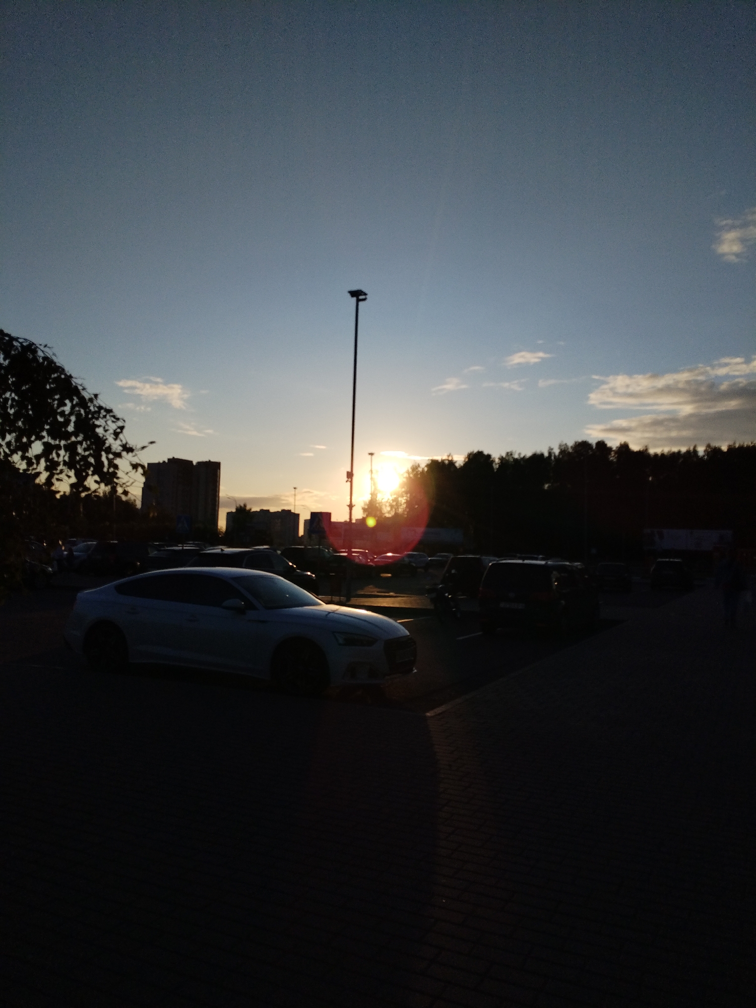 The image captures a street view in Mahilyow, Belarus, during sunset. The sky is a gradient of blue and orange, with scattered clouds. The sun is low on the horizon, casting a warm glow and creating lens flare. In the foreground, a white car is parked on a paved surface. Several other cars and buildings are visible in the background, silhouetted against the setting sun. A tall lamppost stands prominently on the right side of the image. Trees line the horizon, forming a dark backdrop to the scene. The overall impression is one of a peaceful evening in an urban setting.