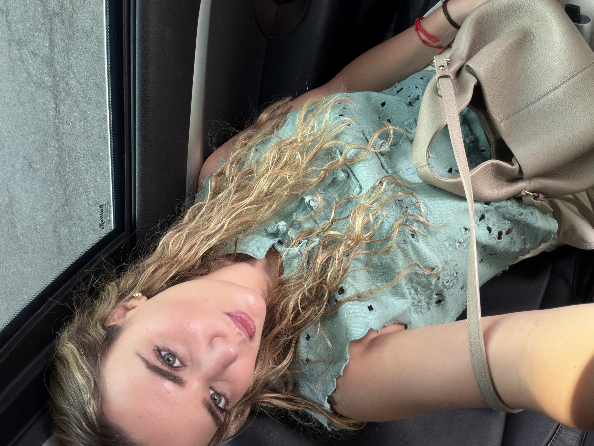 A woman is lying down, possibly in a car, inside an environment in the city of Unknown, Unknown. The woman has light skin, long curly blonde hair, and is wearing a light green cut-out top. A beige handbag rests near her. The background features a car window and dark interior.