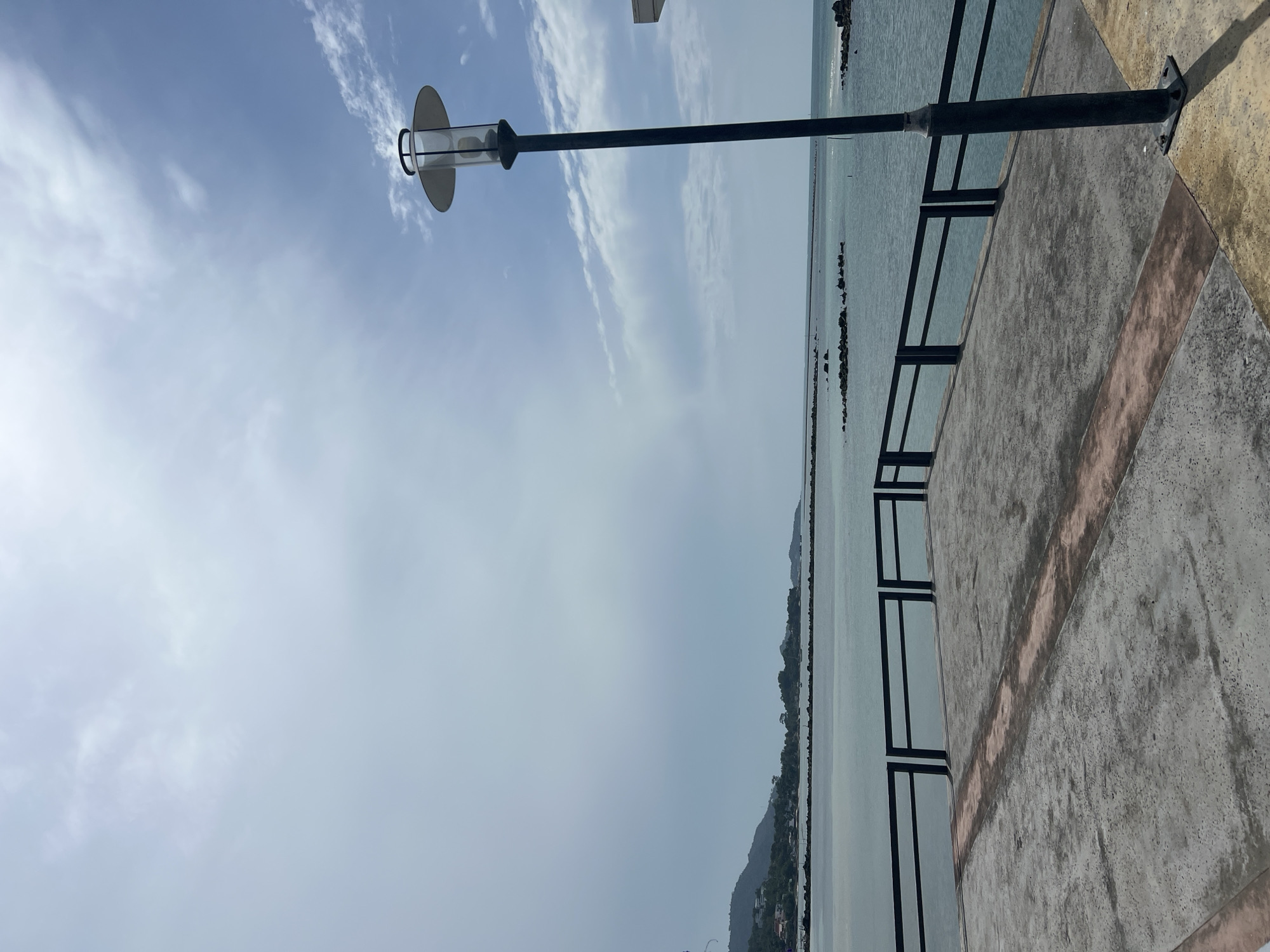 A waterfront promenade in the city of Ko Samui District, Thailand, features a concrete walkway with a dark metal railing overlooking the sea. A tall black lamp post with a circular light fixture stands on the right side of the walkway. In the distance, a calm body of water separates the promenade from a lush, green coastline with scattered buildings. The sky is partly cloudy, with a mix of bright blue and white clouds.