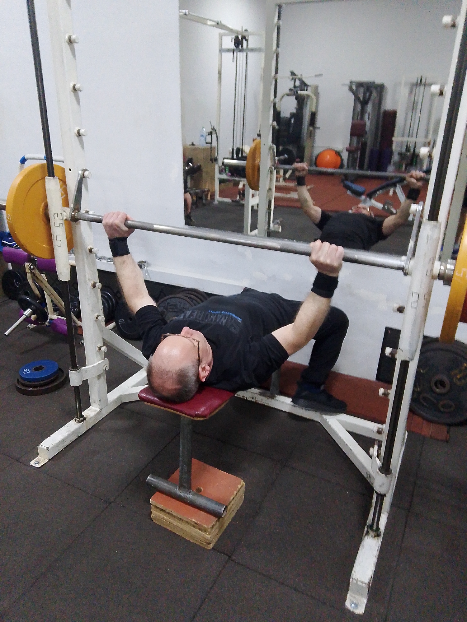 The image shows a man in a gym in Chaikovsky, Russia. The man is lying on a weight bench, performing a bench press using a Smith machine. He is wearing a black shirt, black pants, and black wrist wraps. He has glasses and a bald head. Two yellow weight plates are on each side of the barbell. A mirror behind the man reflects the scene. The gym has rubber flooring. In the background, other gym equipment is visible.