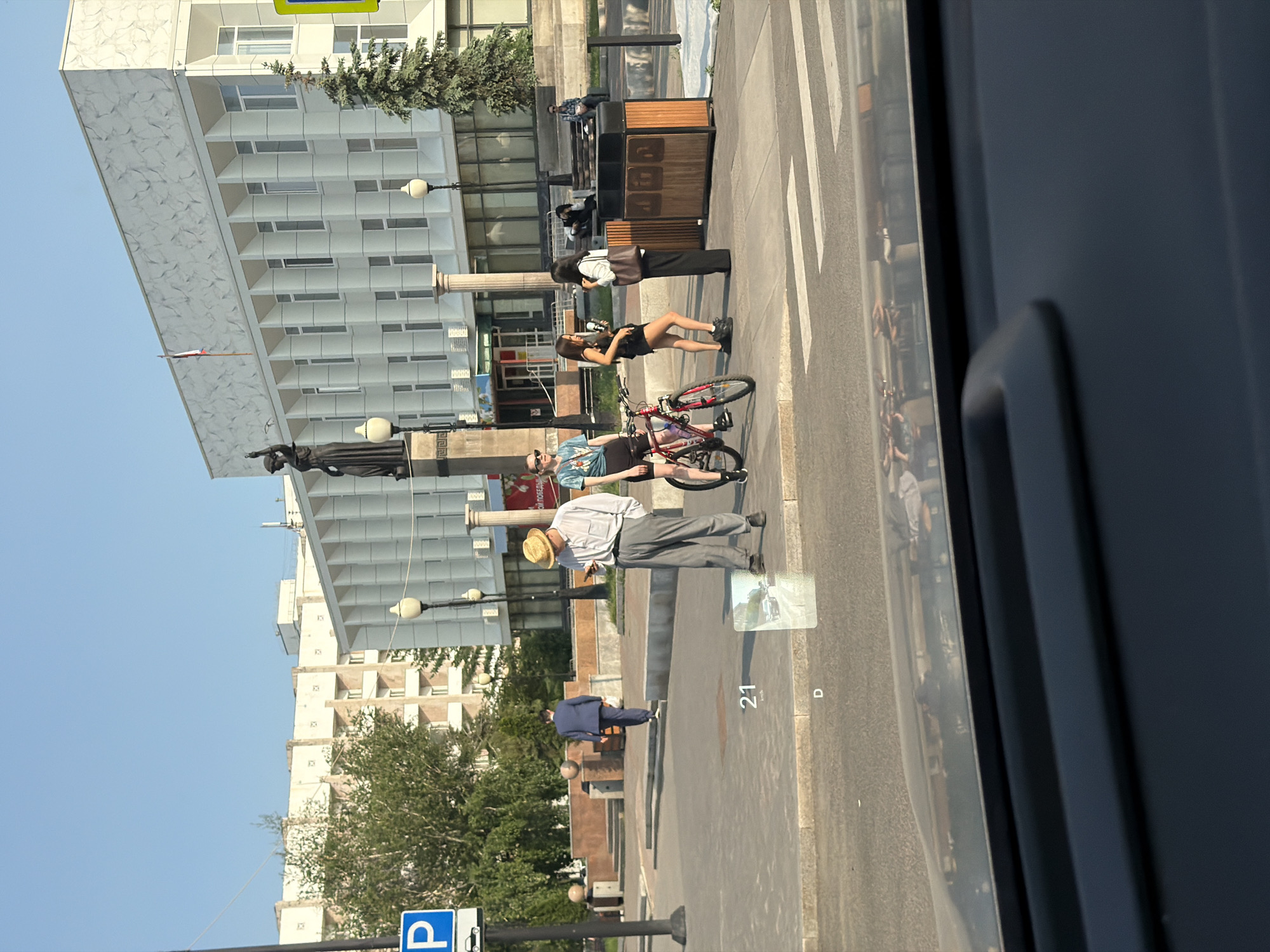 The image captures a street scene in front of a large, modern, white building with a flag on its facade. Several people are visible on the sidewalk. To the left, a man in a straw hat and light-colored pants is looking at his phone. A red bicycle is next to a young woman wearing a t-shirt and shorts. Another woman, dressed in black, stands nearby. Further to the right, two women are engaged in conversation, one holding a coffee cup. In the background, a man in a blue suit walks away from the camera. The environment includes a statue of a woman with a raised arm, a parking sign, trees, and streetlights. The scene appears to be a public space in the city of Krasnoyarsk, Russia, possibly near an administrative building given the architecture. A digital display in the foreground shows "21" km/h and "D," indicating it is likely from a vehicle's heads-up display.