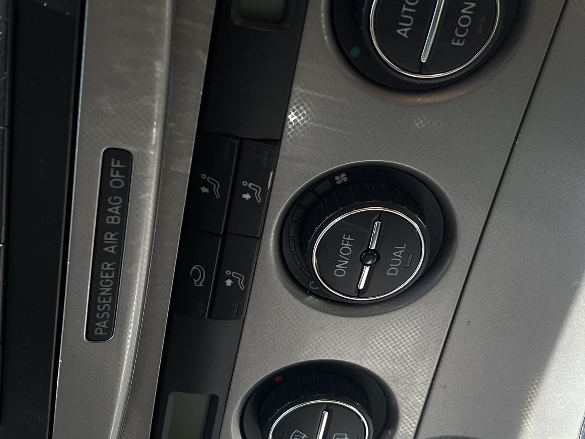 This media file is a close-up, vertically oriented shot of a car's climate control panel. The subjects are the various buttons, knobs, and a label on the dashboard. The panel is made of a textured gray plastic with some metallic accents.
Visible objects include:
- A label that reads "PASSENGER AIR BAG OFF".
- Several buttons with icons: one appears to be a fan with arrows indicating airflow direction, and another with a recirculation symbol.
- Two rotary knobs with chrome rings. The top knob has markings for "ON/OFF" and "DUAL". The bottom knob has symbols that are partially obscured but appear to be related to temperature or fan speed. There is a small red indicator light illuminated on the bottom knob.
- Part of a larger dial with text that is partially visible, showing "AUTO" and "ECON".
The scene is a car interior, specifically focusing on the central console where climate controls are located. There are no people visible. The lighting suggests it could be daytime, as there is a bright reflection on the textured plastic, but the specific time of day or weather conditions are not discernible. The location cue of "Lviv, Ukraine" is provided separately and is not visible in the image itself. The activity taking place is not evident from the static image, but the controls suggest the operation of the vehicle's climate system.