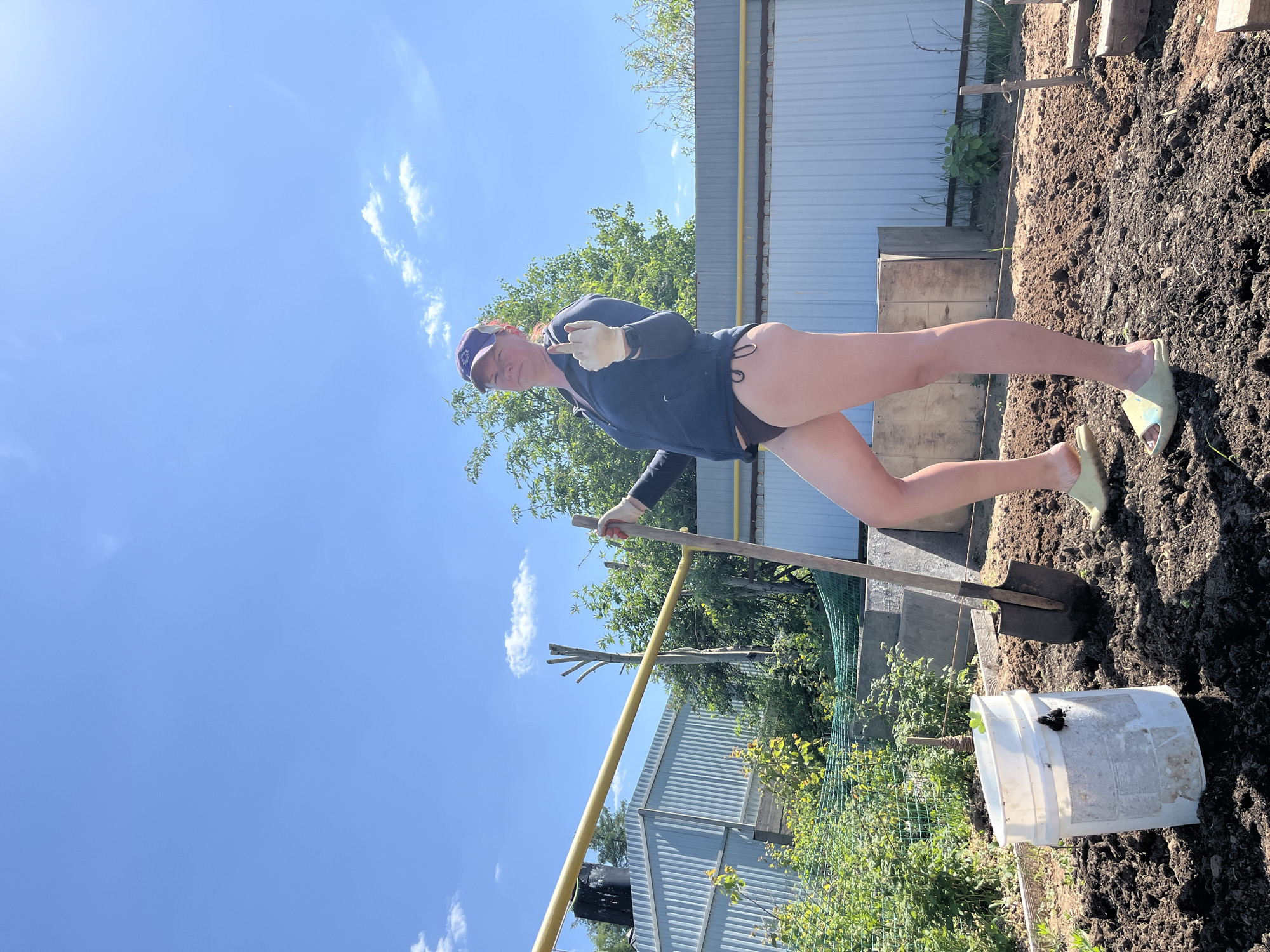 A woman is standing in a garden in what appears to be Chaikovsky, Russia. She wears a dark-colored zip-up shirt, a dark-colored bikini bottom, light-colored gloves, a baseball cap, and light-colored slides. She holds a shovel and gives the finger. A white bucket sits in the garden. A metal fence is in the background.