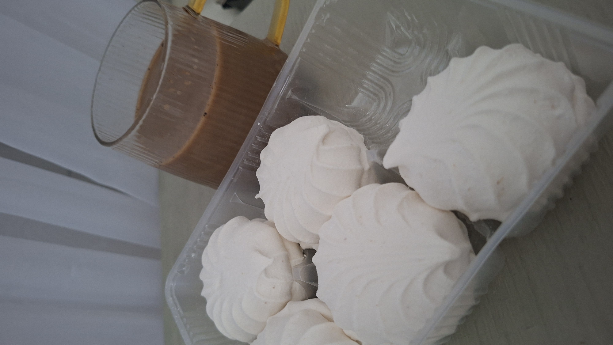A close-up, angled shot shows a clear plastic container filled with five white, fluffy marshmallows, each with a distinctive swirl pattern. To the right and slightly behind the marshmallows, a clear glass mug with vertical ridges contains a brown beverage, possibly coffee or hot chocolate, with a yellow handle. The background consists of white, vertical fabric, likely curtains. The scene is set on a neutral-colored surface, suggesting a domestic indoor environment. There is no indication of the specific location within Myadzyel, Belarus, nor any people or actions.