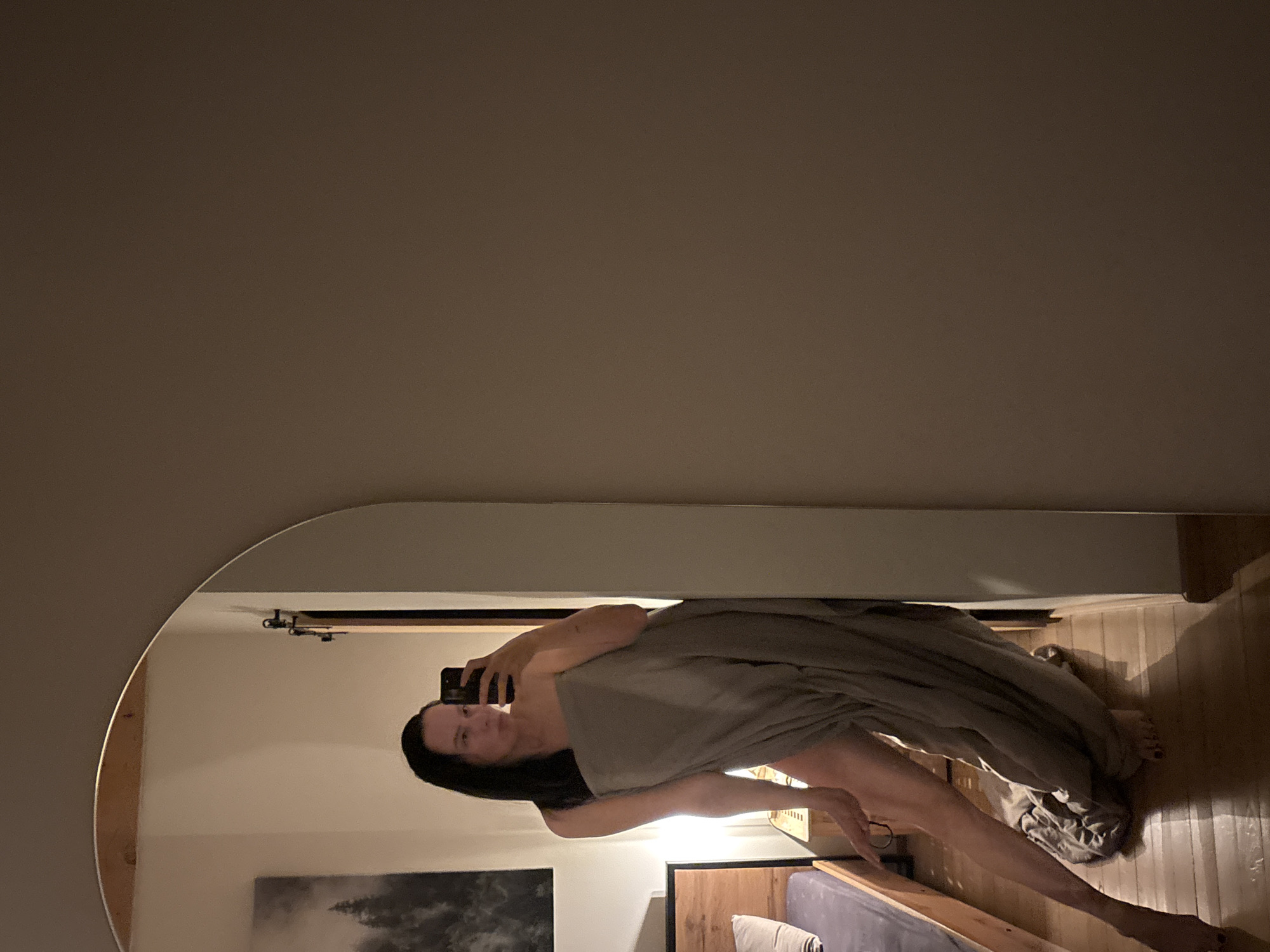 A woman is reflected in a large, rounded mirror. She is holding a phone and appears to be taking a selfie. She is partially covered by a gray blanket or sheet. The background shows a bedroom setting with a bed, artwork on the wall, and wooden floors. The location and city of Chaikovsky, Russia, cannot be confirmed from the visual information provided.