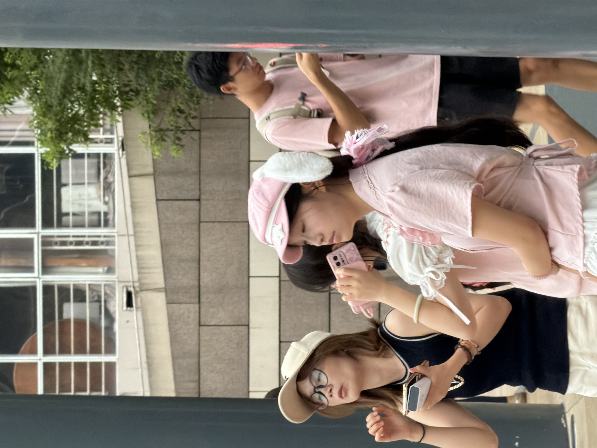In Yuzhong District, China, three individuals are visible outdoors. In the foreground left, a woman wearing a cream-colored baseball cap and glasses, dressed in a navy tank top and white shorts, holds a white rectangular object, possibly a portable charger, in her left hand. Beside her to the right, a young girl in a pink dress with white ruffled sleeves and a pink hat adorned with white fluffy ears looks at a pink phone. Behind her, a young man in a pink t-shirt and black shorts, wearing glasses and a backpack, looks down at his phone. The background features a brick wall and a building with windows.