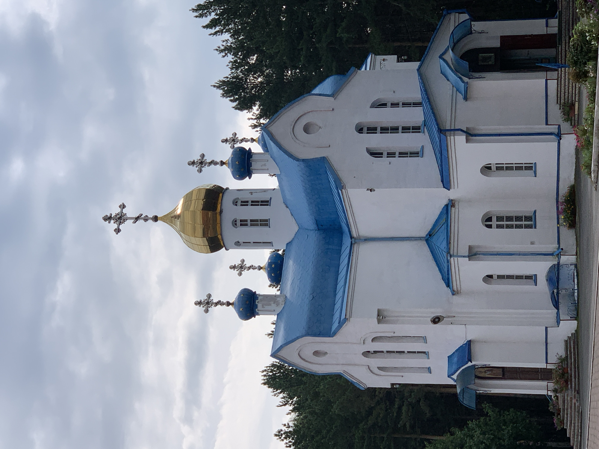 An Orthodox church with white walls and a bright blue roof stands prominently in a forest clearing. The church features multiple domes topped with crosses, the largest being a prominent golden dome. The architectural style includes arched windows and entrances with blue canopies. Dense evergreen trees form a backdrop to the building, suggesting a natural or semi-rural setting. The sky above is overcast with grey clouds. In the foreground, there are steps leading to the entrances, adorned with potted flowers. The scene is static, with no people or animals visible. This is the Holy Trinity Church in Prokopyevsk, Russia.