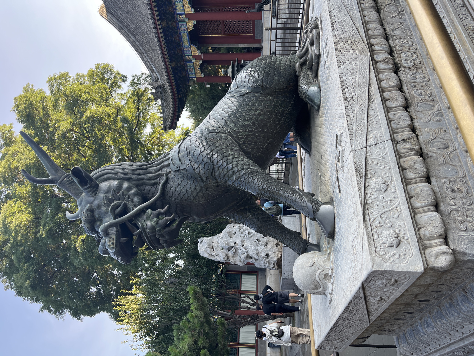 A detailed, medium shot shows a large, weathered bronze statue of a mythical beast, possibly a qilin or dragon, in a courtyard. The statue is dark green and textured, with scales covering its body, a fierce expression, and sharp claws. It is positioned on a decorative stone pedestal with intricate carvings. In the background, several people are visible, some walking and others standing. To the right, a traditional Chinese building with a curved, tiled roof and red columns stands. Trees with lush green foliage are scattered throughout the scene, some partially obscuring the sky. The ground appears to be paved with stones. The lighting suggests it is daytime, with soft shadows.