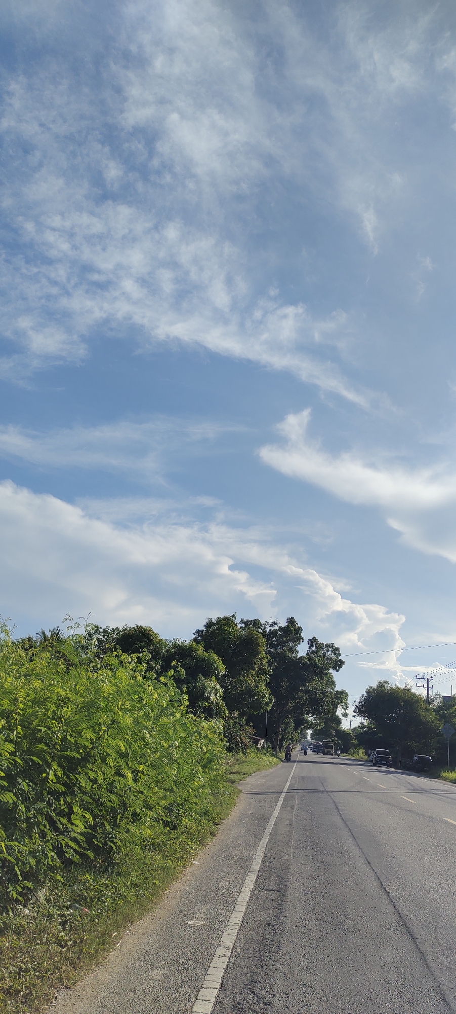 The image depicts a scenic view of a road in Matangpanyang, Indonesia, under a partly cloudy sky. The road, paved with asphalt, stretches into the distance, flanked by lush green vegetation on the left and a line of trees on the right. Several vehicles, including cars and a bus, are visible on the road. The sky is a vibrant blue, adorned with scattered white clouds that create a dynamic and atmospheric effect. The overall impression is one of a peaceful rural landscape on a bright day.