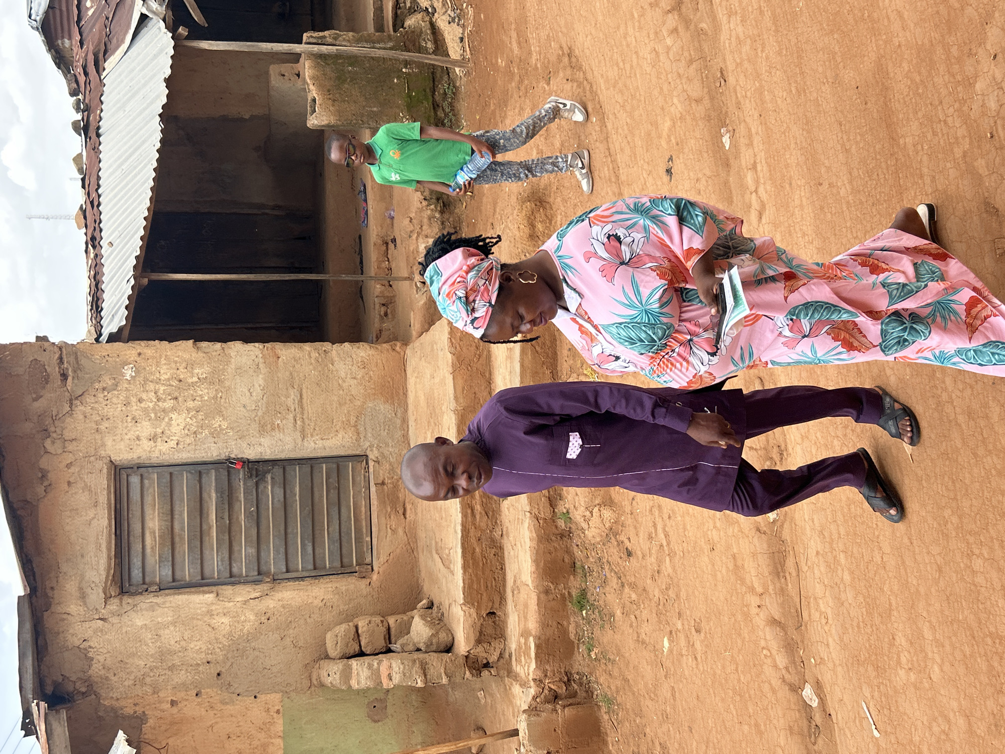 The image depicts three individuals in an outdoor setting within the city of Jos, Nigeria. The foreground features a man wearing a purple long-sleeved shirt and matching pants, and black sandals. Behind him is a woman wearing a pink and white floral patterned dress and matching head wrap, who is looking down at a small object in her hands. A boy in a green shirt and camouflage pants stands further back, holding a plastic water bottle. The background consists of a weathered building with a corrugated metal roof. A window with metal bars is visible. The ground is reddish-brown earth.