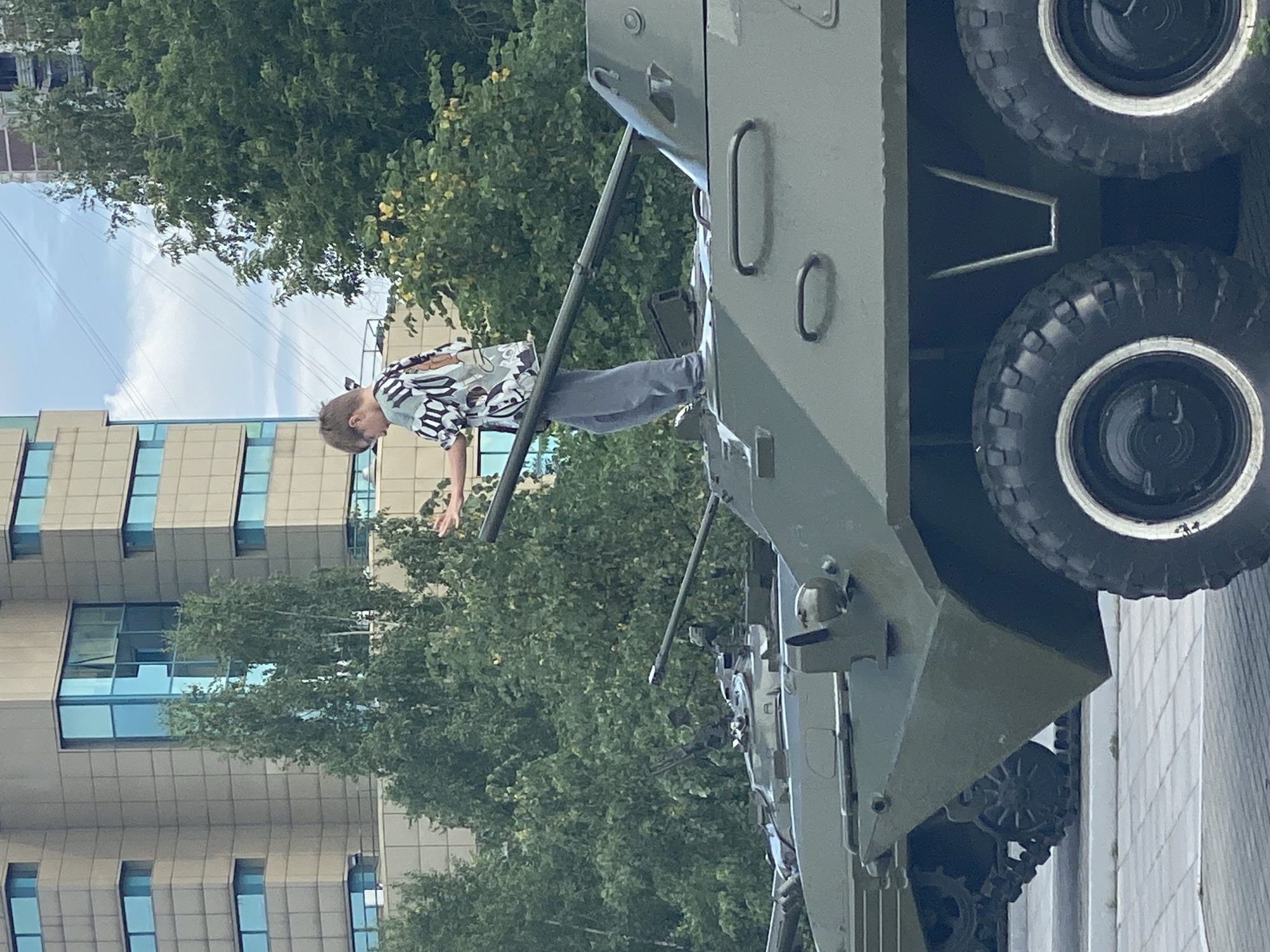 A young boy stands on top of a green armored personnel carrier, his left hand outstretched. He wears a short-sleeved shirt with a black and white abstract pattern and grey pants. The APC is part of a display, with another APC visible behind it. In the background, a modern building with blue-tinted windows rises above a dense canopy of green trees. The scene appears to be outdoors, possibly in a city park or public square in Novokuznetsk, Russia, given the architectural style of the building and the type of military vehicles on display. The sky is partly cloudy.