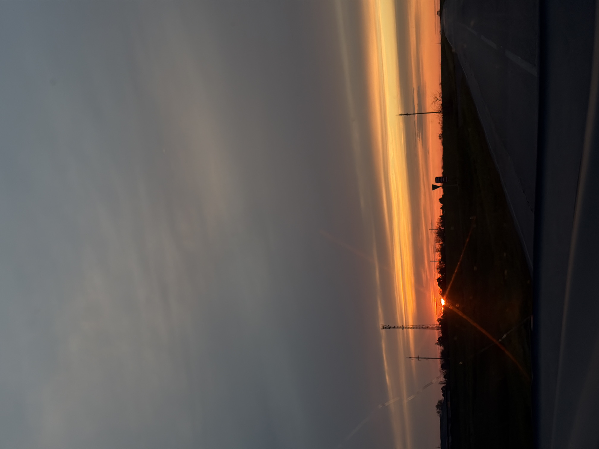 The image captures a vibrant sunset or sunrise over a semi-rural landscape, viewed from the interior of a moving vehicle, likely in Alexandrovka, Russia.
The sky dominates the upper two-thirds of the frame, showcasing a spectacular display of warm colors. Layers of clouds are illuminated in brilliant shades of orange, deep yellow, and red near the horizon, gradually transitioning to softer grays and blues higher up. The sun, a bright, intense orb, is visible in the lower right quadrant, partially obscured by the distant landscape, casting strong rays and creating noticeable lens flare effects.
Along the horizon, a dark silhouette defines the land, featuring sparse trees, bushes, and various man-made structures. Prominent among these are utility poles and at least one tall communication or cellular tower, standing starkly against the glowing sky. A straight road, likely an asphalt highway, occupies the right side of the image, with a white dashed line marking the lane, extending into the distance. The dark, curved edge of a vehicle's interior is visible on the far right, framing the scene.
The time of day is clearly sunset or sunrise, characterized by the low position of the sun and the dramatic, colorful sky. The weather appears clear with scattered clouds. There is no visible text in the image. The scene evokes a sense of tranquility and the natural beauty of the day's beginning or end.