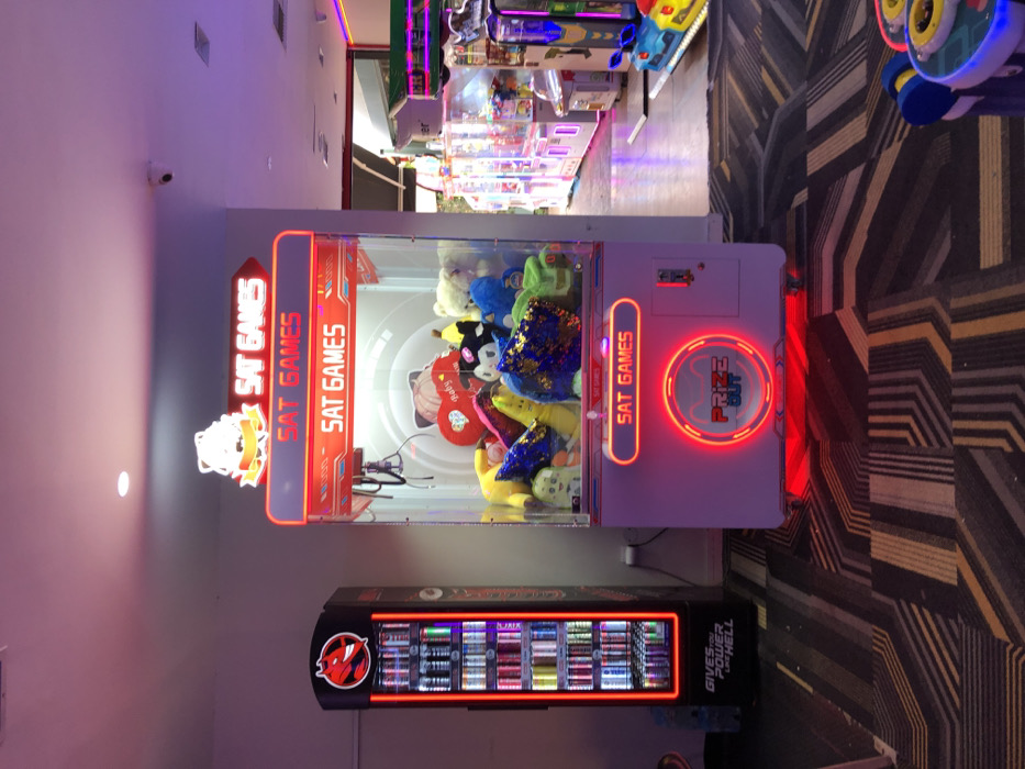 This image presents an indoor scene within an arcade or entertainment center in Iraklio, Greece. The main subject is a large, white claw machine, brightly illuminated with red neon lights, standing prominently in the foreground. Inside the machine, a variety of plush toys are visible, including a black and white panda-like figure, a yellow character, a blue creature, and a red heart-shaped pillow, which has the word "Baby" on it. The text "SAT GAMES" is displayed multiple times in red neon on the machine's front and sides. A circular red neon outline highlights a section labeled "PRIZE OUT."
To the left and slightly below the claw machine, a tall, black vending machine stands. It features internal illumination showcasing various items, likely snacks or beverages, and has a distinctive red devil-like logo. Text on the side of this vending machine reads "GIVES YOU POWER LIVE HELL." In the background, beyond the claw machine, other arcade games with colorful lights can be glimpsed, hinting at a wider amusement environment. The floor is covered with a geometric patterned carpet in shades of brown, black, and beige. The lighting is entirely artificial, dominated by the vibrant red neon of the game machines, creating an inviting and dynamic atmosphere. No people are visible in the frame, suggesting the machines are ready for play.
