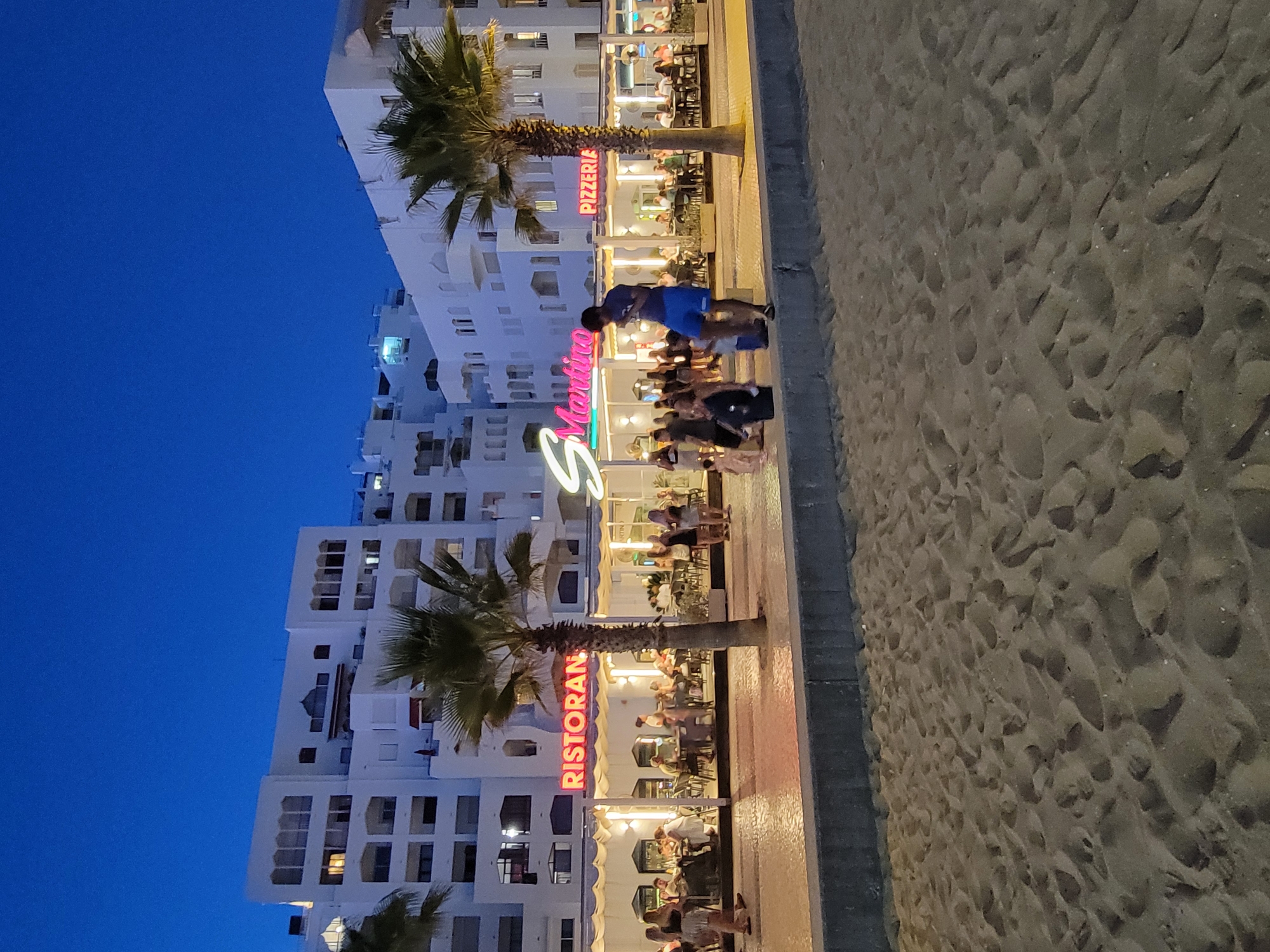 In Quarteira, Portugal, at dusk, a beachfront promenade features the illuminated signs for "Ristoran" and "Pizzeria," adjacent to a restaurant named "S Martino." White buildings with multiple balconies line the background, partially obscured by palm trees. A sand-covered area dominates the foreground. Several people are visible, including a man wearing blue shorts and a dark shirt standing on a low wall, and other individuals dining at tables and walking along the promenade. Children are also present, one appearing to be carried by an adult. The scene captures a lively evening atmosphere by the sea.