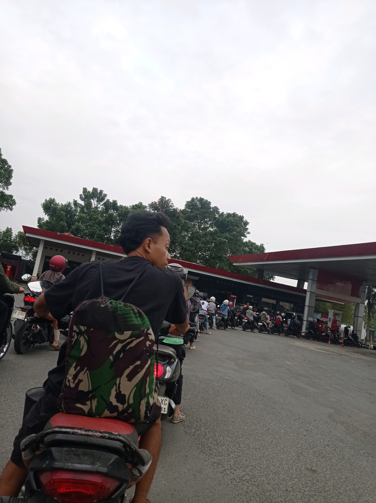 In Bojong, Indonesia, a group of motorcyclists waits in line at a gas station. The image focuses on a man in the foreground, wearing a black t-shirt and a camouflage backpack, as he sits on his motorcycle. Other motorcyclists, many wearing helmets, are visible in the background, forming a queue under the gas station's canopy. The sky is overcast, and trees line the perimeter of the gas station. The overall scene depicts a common sight of people waiting to refuel their vehicles.
