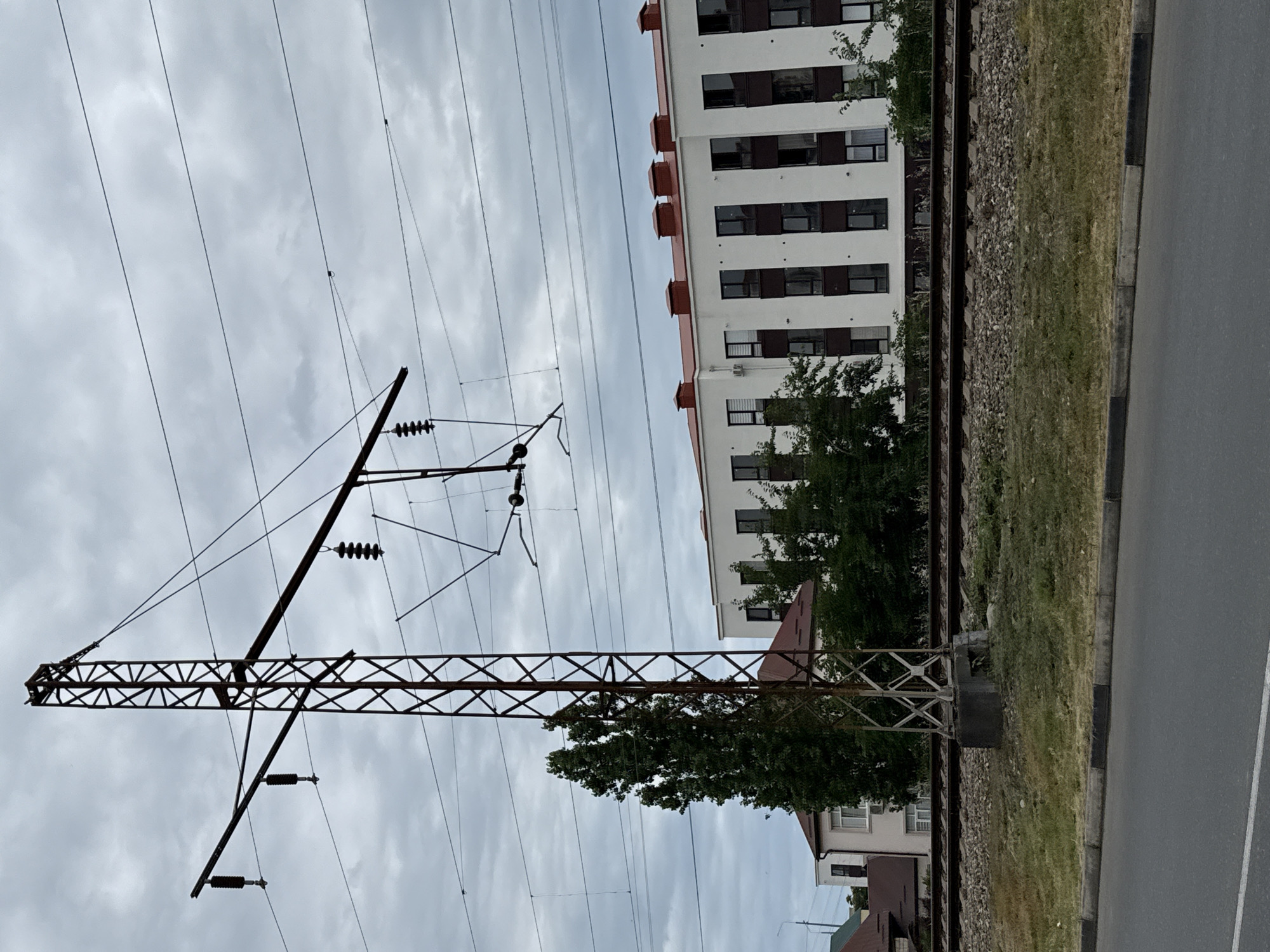 The image depicts a cloudy day in Derbent, Russia, with a focus on railway infrastructure and urban development. In the foreground, a paved road with a curb runs horizontally, bordered by a grassy verge. Behind the verge, railroad tracks are visible, with ballast stones and adjacent vegetation. A tall, metal lattice power pole stands prominently near the tracks, supporting overhead electrical wires for a railway system. The pole is weathered and shows signs of rust.
In the background, a modern, multi-story apartment building with a red-tiled roof and white facade dominates the scene. The building features numerous windows with dark frames and a row of distinctive chimney-like structures along the roofline. To the left of the apartment building, a tall, leafy tree is situated, partially obscuring a smaller, darker-roofed residential building. The overall environment suggests an area where urban expansion meets transportation networks, under a overcast sky. No people or animals are visible in the frame.