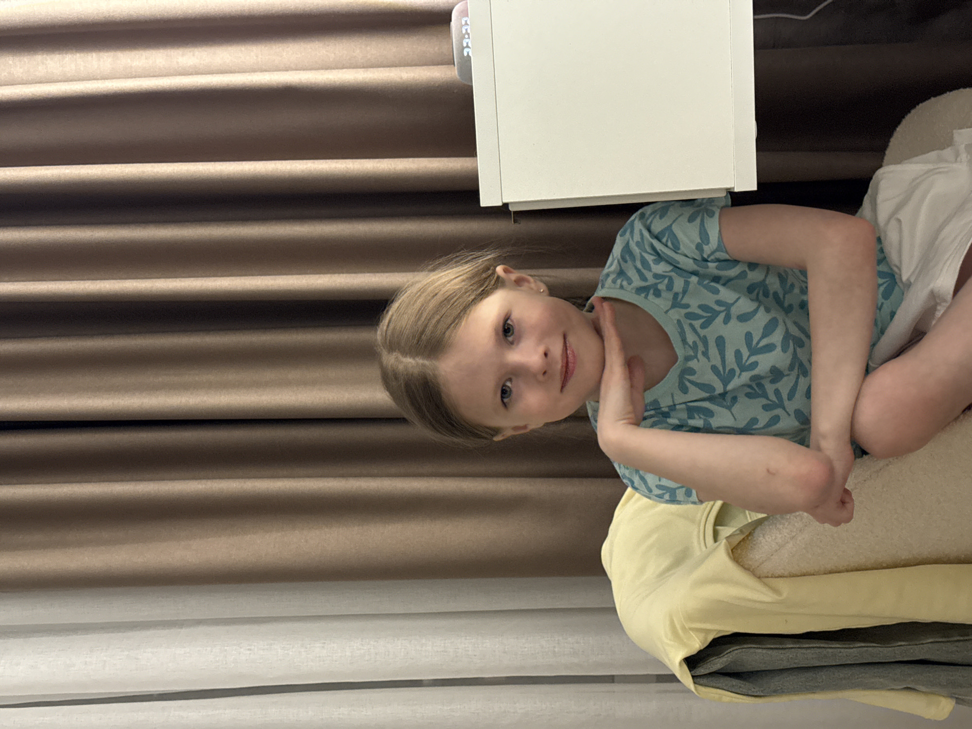 A young girl is sitting, possibly on a couch or bed, in what appears to be an indoor setting. The background features brown horizontal drapes. A white cabinet or table is positioned to the upper-right of the girl. The girl has blonde hair, blue eyes, and is wearing a blue shirt with a pattern. Her posture includes one hand raised near her face. The photo's orientation is rotated, with the background running vertically along the left side of the frame. It is not possible to determine if the photo was taken in Izhevsk, Russia.