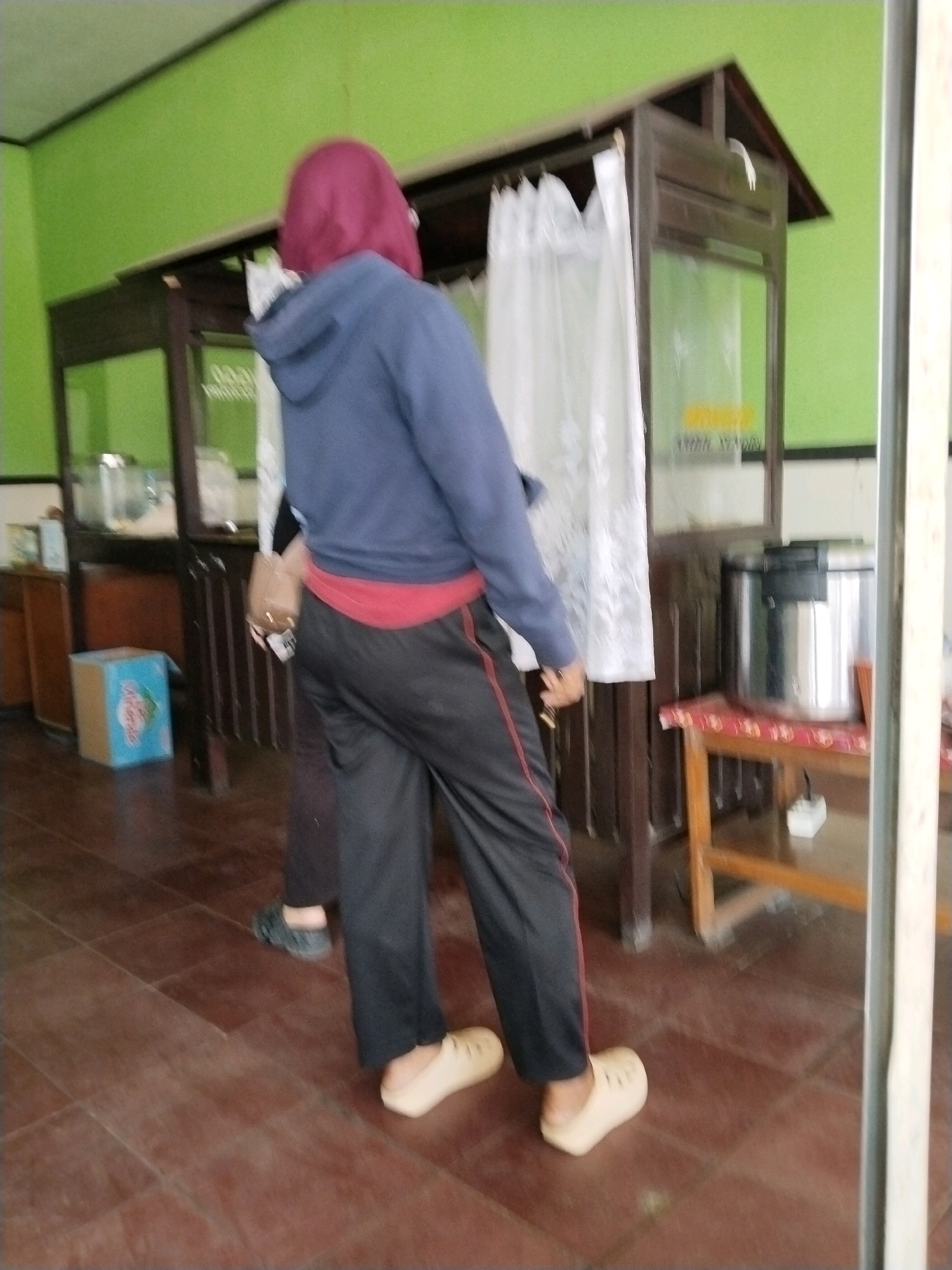 In Mergayu, Indonesia, a person wearing a purple hijab, a blue hoodie, a red sash, and black pants with red stripes stands facing away from the camera. They are wearing light-colored clogs. Another person, whose lower legs are visible, is wearing dark shoes and dark pants. The scene takes place indoors, with a green wall in the background. A wooden structure with white curtains is prominent, and a stainless steel container sits on a wooden table to the right. A blue and white box is on the floor to the left. The floor is tiled with reddish-brown tiles.