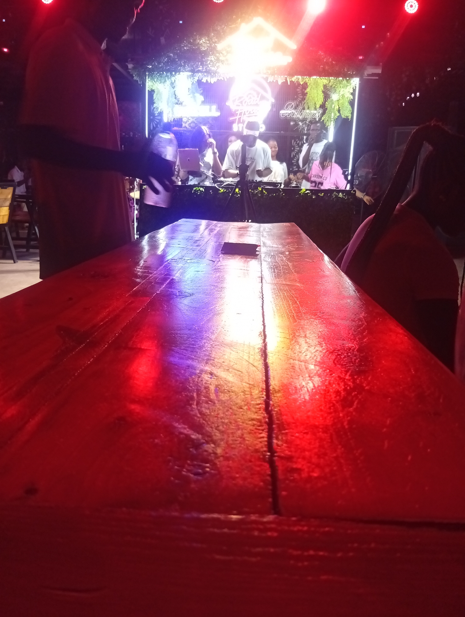 The image depicts a lively night scene at an event space, likely a bar or club, in Howayi, Nigeria. The foreground is dominated by a long, glossy wooden table, highly reflective of the vibrant red and purple artificial lights illuminating the space. The table appears wet or has a highly polished surface, catching the light in streaks.
On the left side of the frame, a person in a short-sleeved, light-colored shirt stands partially silhouetted, their arm extended towards the table, suggesting they might be serving or interacting with an object on the table. In the background, behind the table, a DJ booth is set up. Several people are visible around this area. A DJ, wearing a white t-shirt and a cap, is positioned behind the equipment. Other individuals are present, including one person in a pink t-shirt with "CARSON CO" visible on it, and another in a white t-shirt. Artificial foliage decorates the area above the DJ booth.
The setting is characterized by dynamic, colorful lighting, with red spotlights and neon-like signs creating a festive atmosphere. Partially visible neon text above the DJ booth includes words such as "Road" and "Ballroom." The overall activity suggests a music event or party in full swing, with people gathered to enjoy the entertainment. On the far right, a person with dark braided hair is partially visible, facing away from the camera. The time of day is clearly night, indicated by the dependence on artificial lighting.