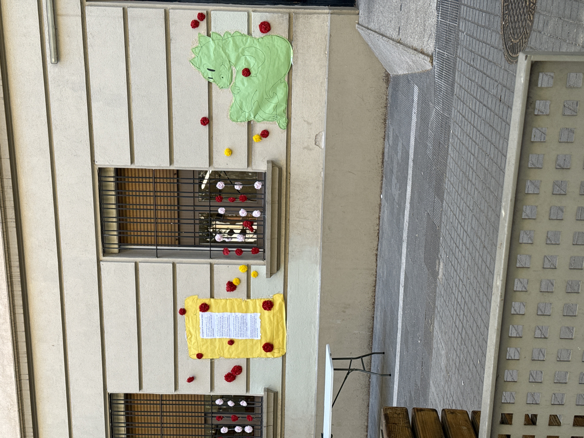 The image depicts a building facade in Barcelona, Spain, adorned with decorations. The facade is characterized by alternating horizontal segments and vertical architectural elements. The decorations consist of a green dinosaur cutout, a yellow fabric sheet with text, and numerous pom-poms in red, yellow, white, and pink. These adornments are affixed to the facade at various points, including above and below window grates. The architectural style is characteristic of the city. Adjoining the building facade is a pedestrian walkway.