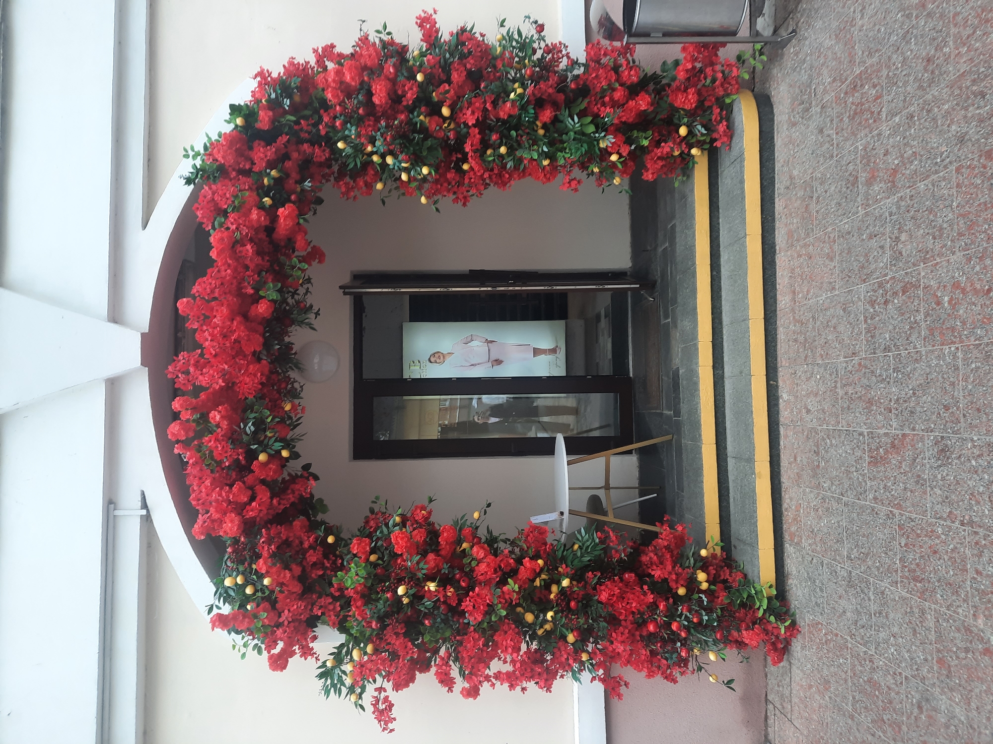 The image shows an arched floral decoration with red flowers and yellow fruit, likely lemons, framing an entrance to a building. The entrance is a double door with glass panels. Inside the entrance, a poster featuring a woman in a light pink suit is visible. To the left of the entrance, there is a small white round table with a chair. The building's facade is light beige, and the area in front of the entrance is paved with grey and red tiles. Two steps with yellow accents lead up to the entrance. The overall scene appears to be an exterior view of a shop or business.