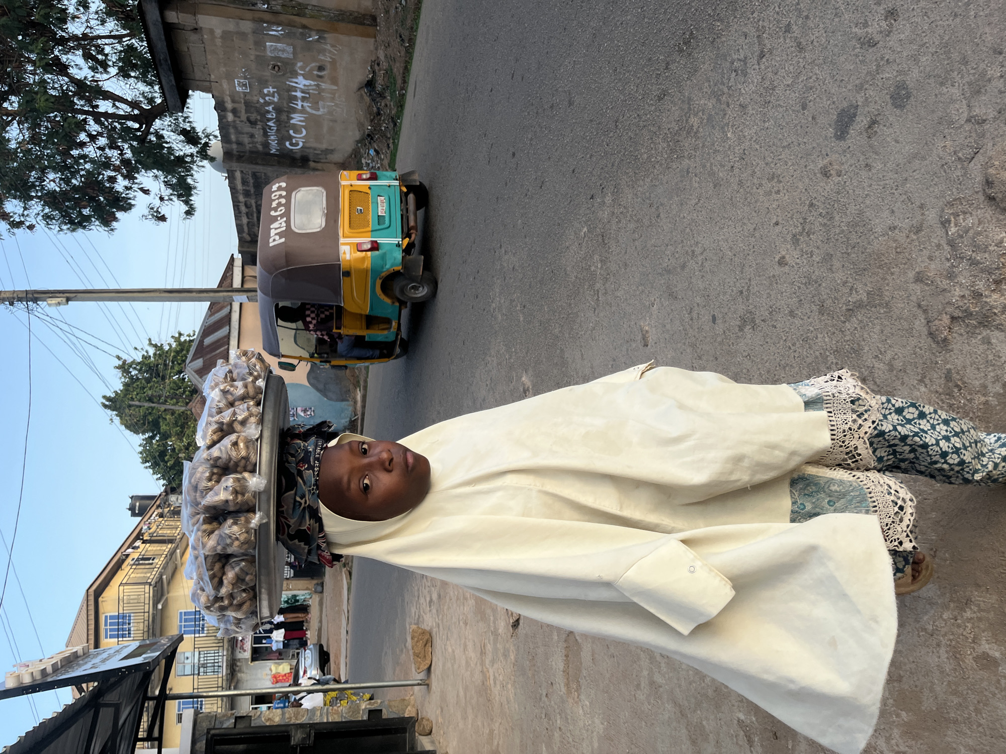 A young woman balances a tray piled high with small, clear plastic bags filled with peanuts on her head, her gaze directed towards the camera. She wears a long, cream-colored garment over patterned fabric, with lace trim visible at her wrists. The setting appears to be a street in Jos, Nigeria, indicated by the presence of a "Keke Napep" auto-rickshaw bearing the license plate "PTA-6393" in the background. The buildings and infrastructure are consistent with an urban residential area. There is graffiti on the wall behind the auto-rickshaw. The street is paved, and additional buildings and trees are visible in the background.