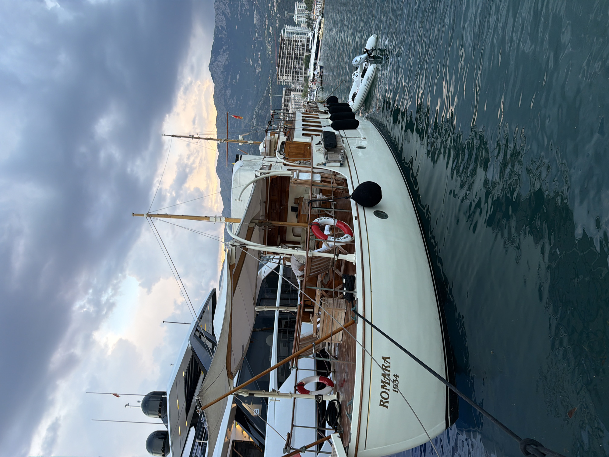 This is a tilted, wide shot of a yacht named "ROMARA 1934" docked in a harbor. The yacht has a classic design with a white hull and wooden accents. Two life preservers are visible on the deck. Behind the yacht, several other boats and buildings are visible, suggesting a marina or waterfront area. In the background, mountains rise above the cityscape, and the sky is filled with dramatic clouds, indicating either sunrise or sunset. The water is calm with gentle ripples. The overall mood is serene and peaceful. Visible text includes "ROMARA 1934" on the hull of the yacht. The location is identified as Budva, Montenegro.