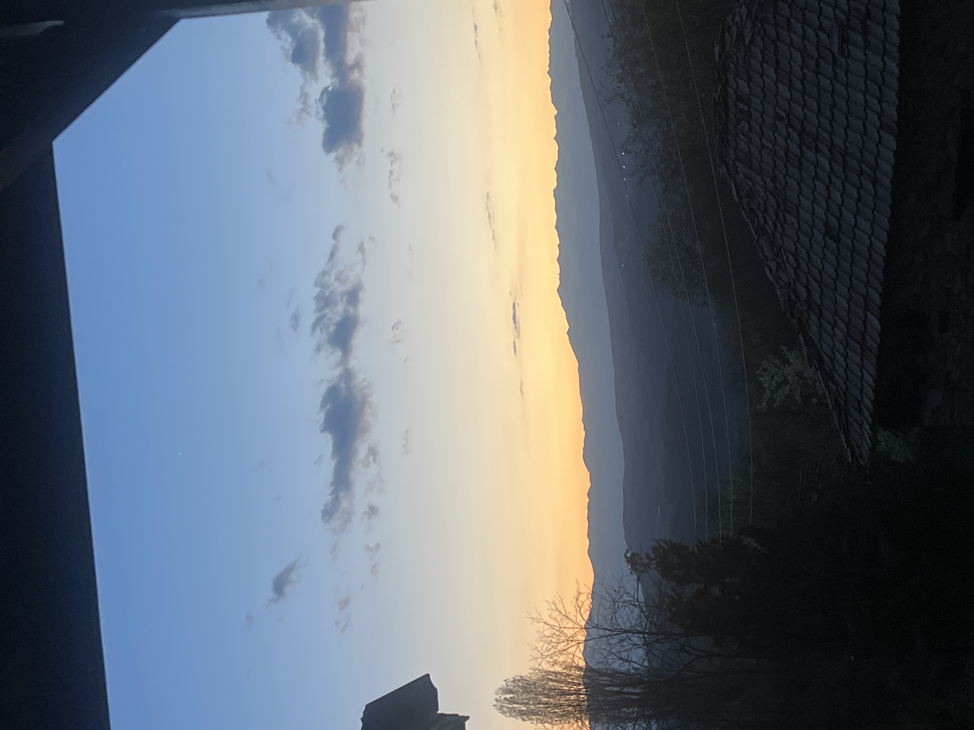 The image shows a landscape view taken from a high angle, likely from inside a building looking out a window. The left side of the image reveals part of the window frame or building structure, which is dark in color. The sky occupies a significant portion of the scene, transitioning from blue with scattered clouds on the left to a gradient of orange and yellow near the horizon. The horizon is lined by a mountain range, appearing in shades of gray and blue. In the foreground, there are silhouetted trees with bare branches. To the right is a building or structure with a dark, textured roof. Powerlines are strung across the mountainscape. The specific location in Kribul, Bulgaria cannot be determined from the image alone without additional information.