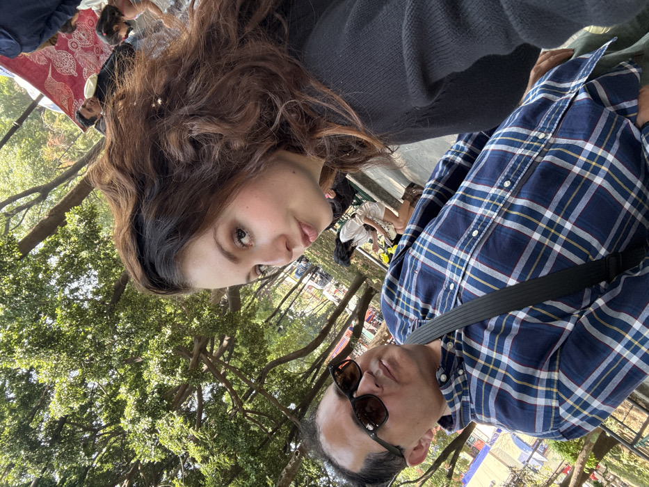 The image presents two individuals in the foreground of an outdoor, tree-lined public space, consistent with a park or market area within Mexico City, Mexico. In the upper right quadrant, a female with long, voluminous brown hair and a dark-colored top is seen looking directly towards the camera, her lips slightly puckered. To her left, occupying the lower left quadrant, a male with short, dark hair and dark sunglasses wears a blue and white plaid button-up shirt with a black strap diagonally across his chest; he looks forward with a neutral expression. The background features a dense canopy of green tree foliage under bright daylight. Various individuals are visible in the mid-ground and background: one female in a light-colored garment is bending over near a yellow object, and further back, under a red patterned canopy, additional indistinct figures, including an adult male and a child, are present. The overall environment suggests a lively public area.