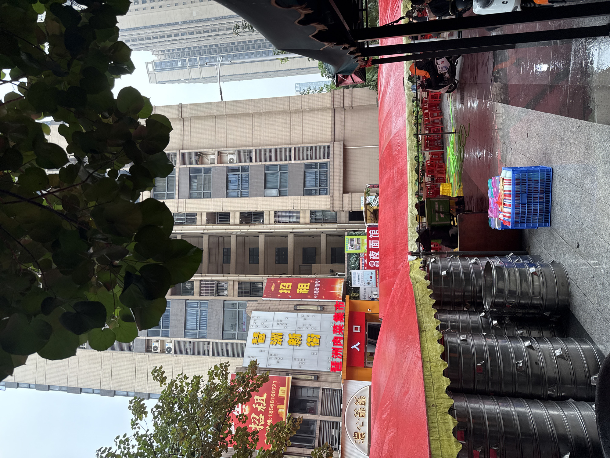The image depicts an outdoor market scene under a red awning in Haizhu District, China. The foreground shows stacks of large, metallic containers, likely for food preparation or storage. Behind them, a red tarp covers a seating area with numerous red chairs and tables, suggesting a restaurant or food stall setup. The ground is wet, indicating recent rain. In the background, several multi-story buildings, characteristic of urban Chinese architecture, rise into a hazy sky. One building prominently displays red and yellow signage with Chinese characters, possibly advertising goods or services. A motorcycle with a rider is parked to the right, and other individuals are partially visible in the distance, engaged in various activities associated with a market. The overall atmosphere is one of a bustling commercial area.