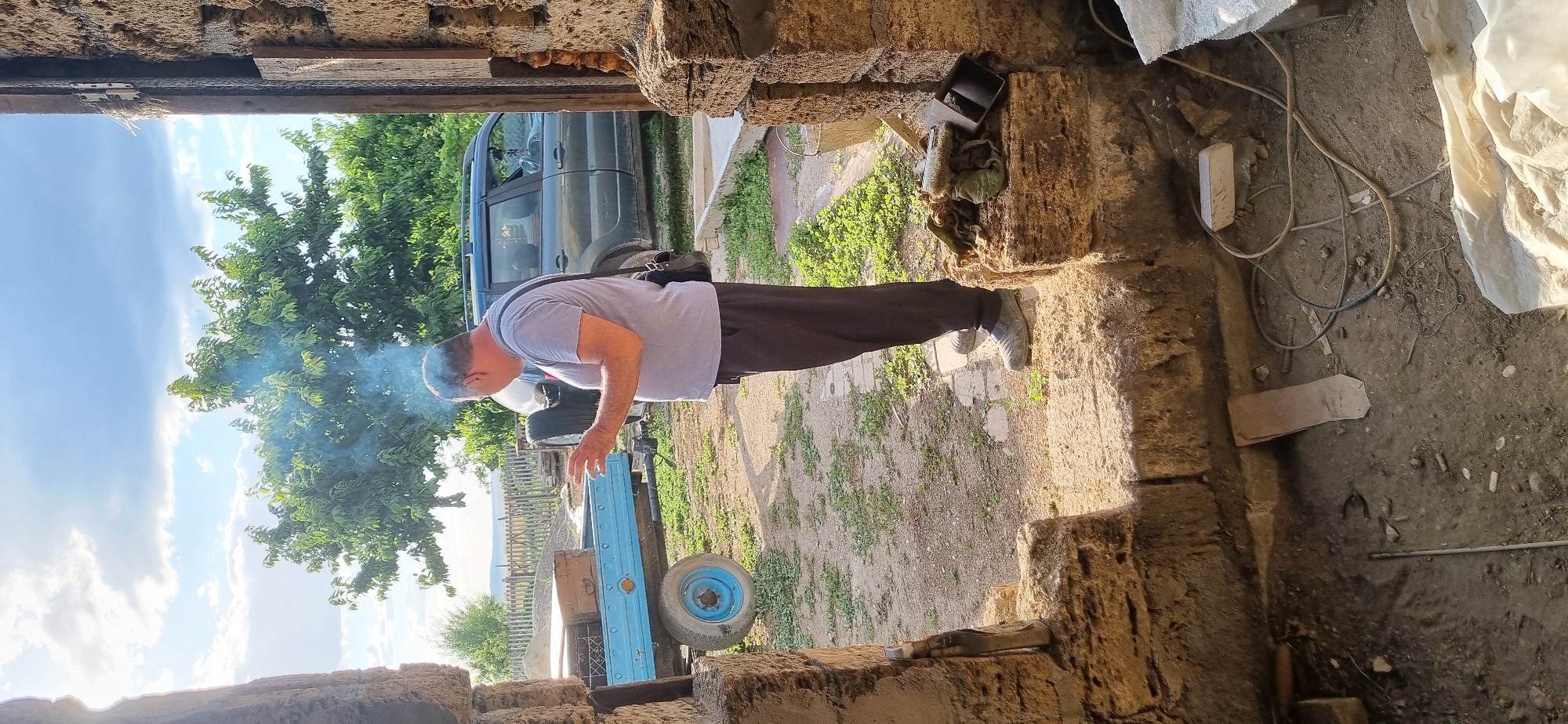 In Blizhnee, Ukraine, an outdoor scene unfolds with a man standing in a partially constructed doorway of a stone building. He is wearing a grey t-shirt and dark pants, with a dark bag slung across his shoulder. The man appears to be smoking, with a plume of smoke rising from his head and dissipating into the bright, partly cloudy sky.
Behind him, a blue trailer is parked, loaded with what appear to be boxes or wooden crates. A dark SUV is also visible further back, along with a spare tire mounted on its rear. The ground is covered with dirt and patchy grass, and scattered debris. To the right of the man, on a stone ledge, are several tools, including a brush and a small container. The foreground shows the rough, unfinished interior of the building, with exposed stone and dirt on the floor, where electrical wires and other construction materials are present. The overall impression is one of a rural or semi-renovated outdoor workspace.