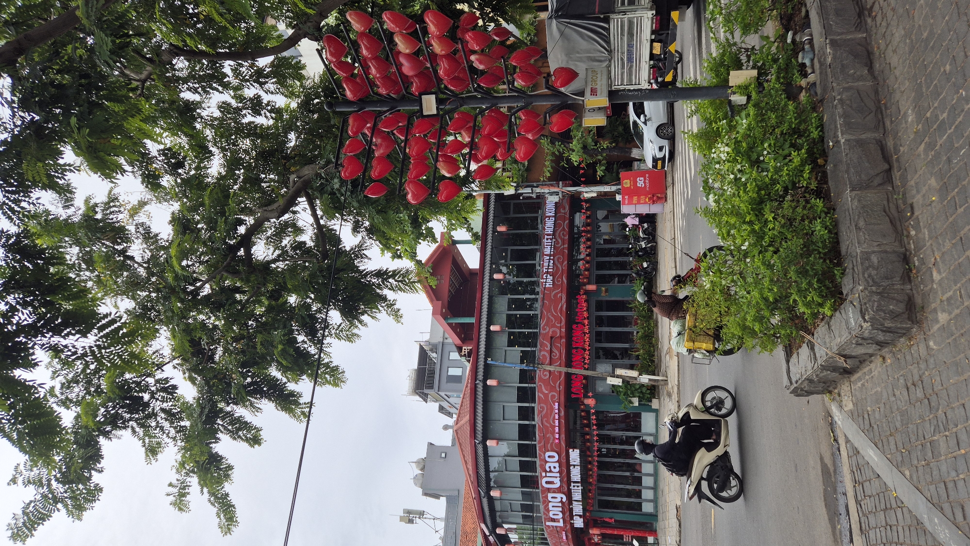 In the city of Đà Nẵng, Vietnam, a street scene unfolds under a cloudy sky. A two-story building with a red tiled roof and large glass windows serves as the backdrop. The building's facade is adorned with red decorative elements and signs in Vietnamese, indicating it is a restaurant named "Long Qiao" and "HAP THUY NHIET HONG KONG." Red lanterns hang along the building's exterior.
In the foreground, a tree-like sculpture decorated with numerous red heart shapes stands prominently on the right side of the image. The street in front of the restaurant is moderately busy. Two individuals on motorcycles are visible: one on a cream-colored scooter on the left, and another on a red scooter carrying a yellow cargo box on the right. A white car and a large truck are also present on the street. Lush green bushes are planted along the curb, separated by a low stone border. The ground is paved with cobblestones.
